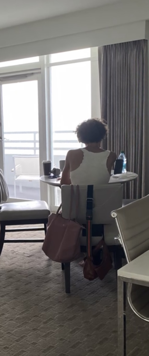 Person sitting at a table near a large window with curtains, holding a water bottle, with a bag on the chair.