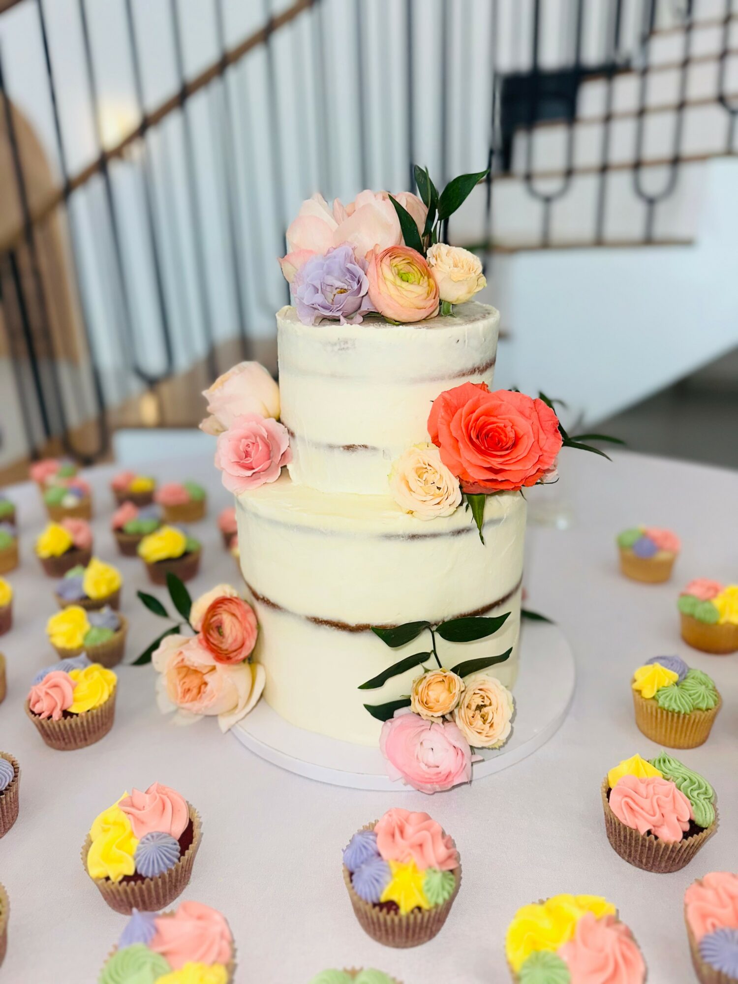 Three-tiered white cake decorated with pink, purple, and orange flowers, surrounded by colorful cupcakes on a table.