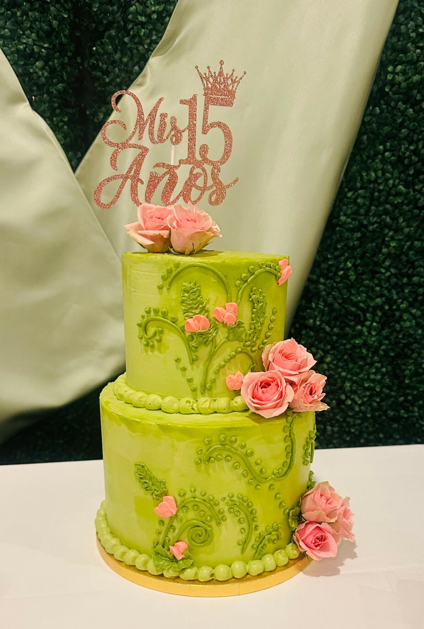 Two-tier green cake decorated with pink roses and a topper reading 'Mis 15 Años' with a crown, celebrating a quinceañera.