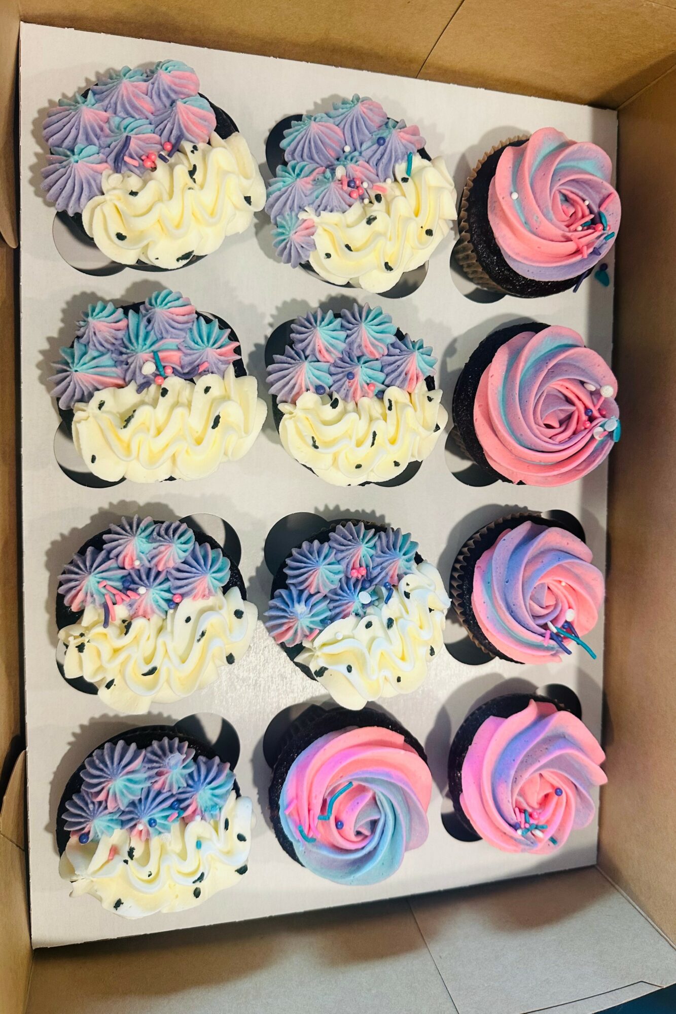 Box of twelve cupcakes with colorful frosting and sprinkles, arranged in three rows of four, with pink and blue swirls.