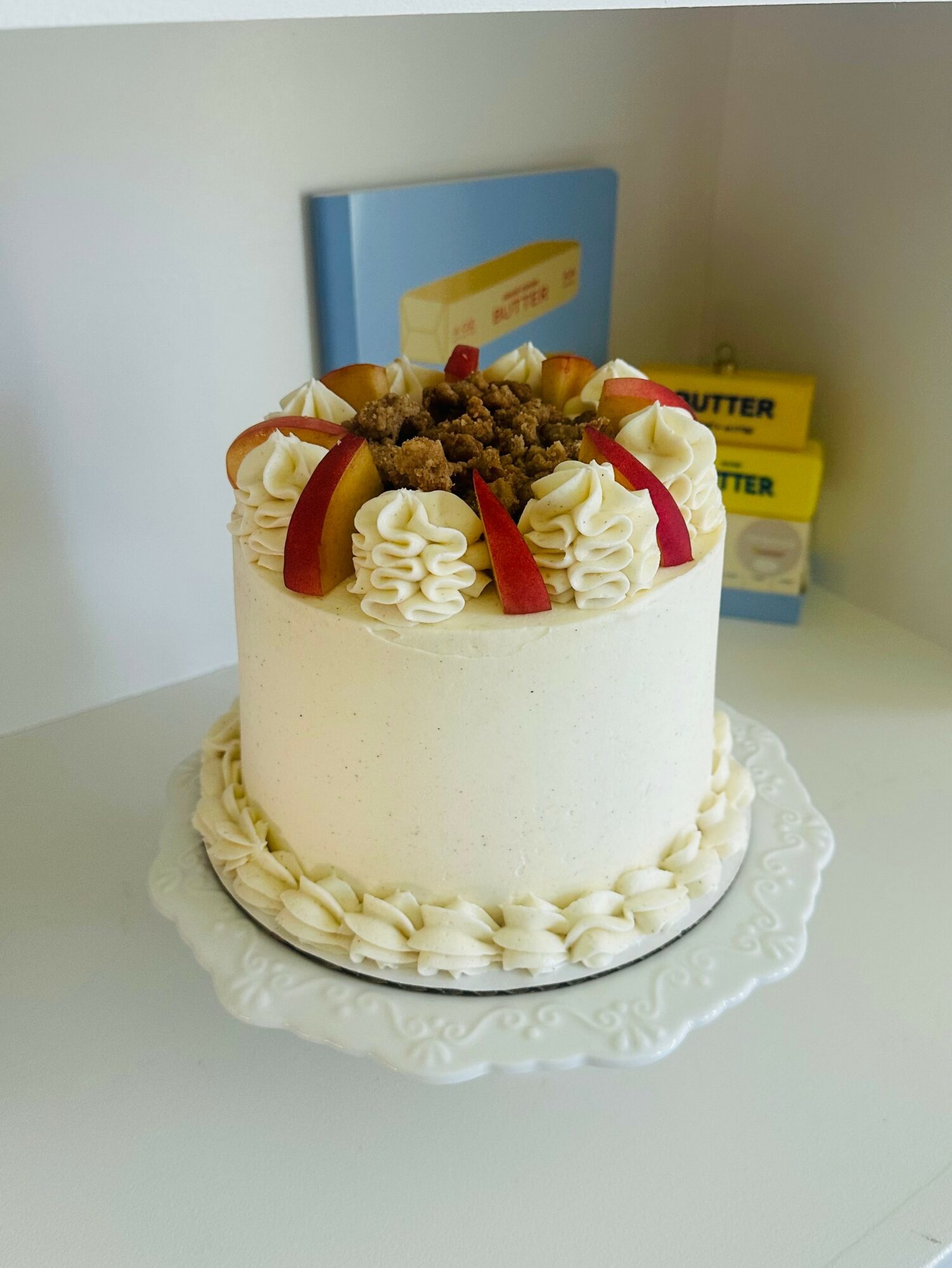Round cake with white frosting, topped with whipped cream, fruit, and chocolate, on a white surface with books in background.