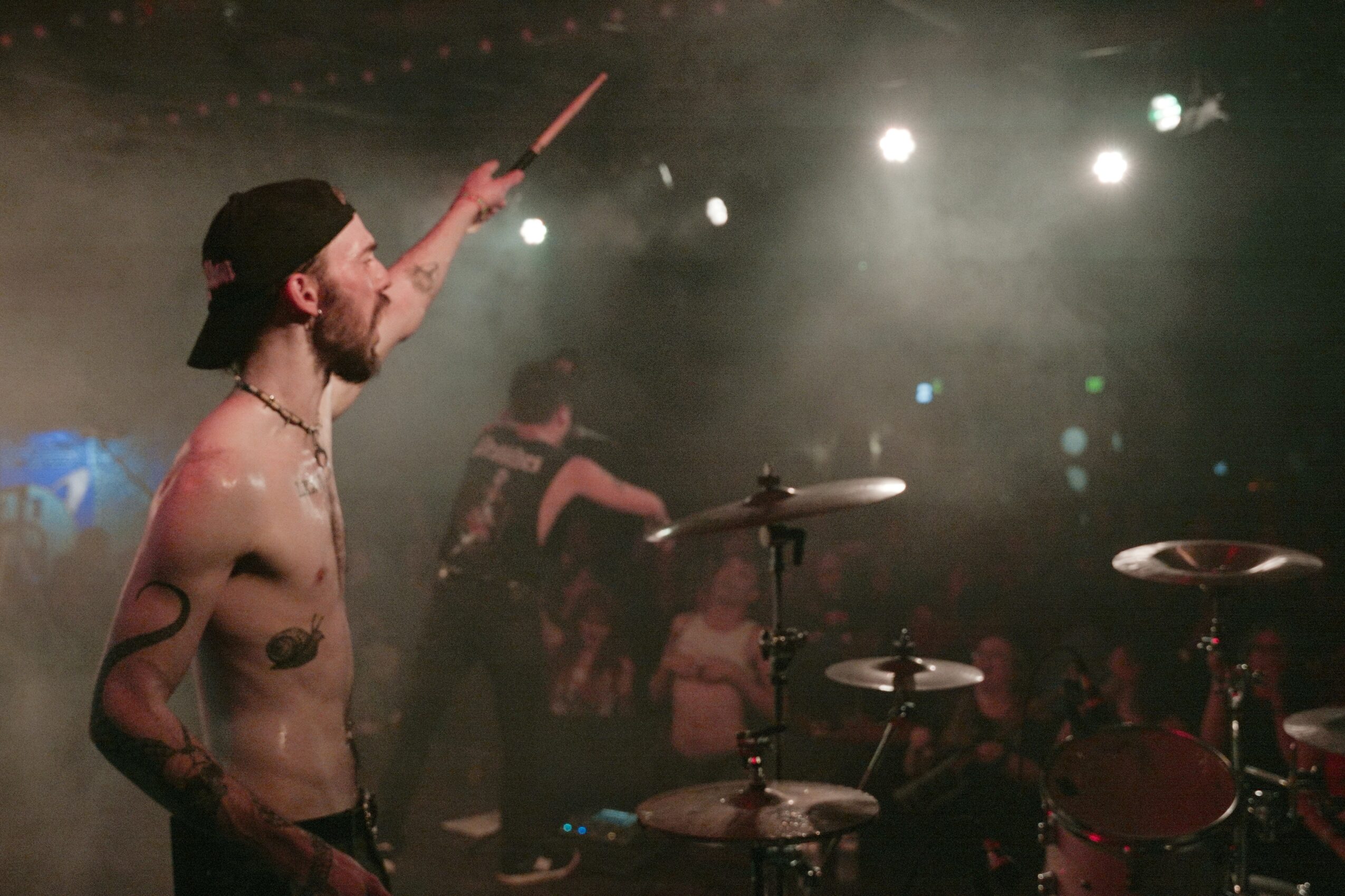 A shirtless drummer with a beard and cap plays on stage, with a crowd and stage lights in the background.