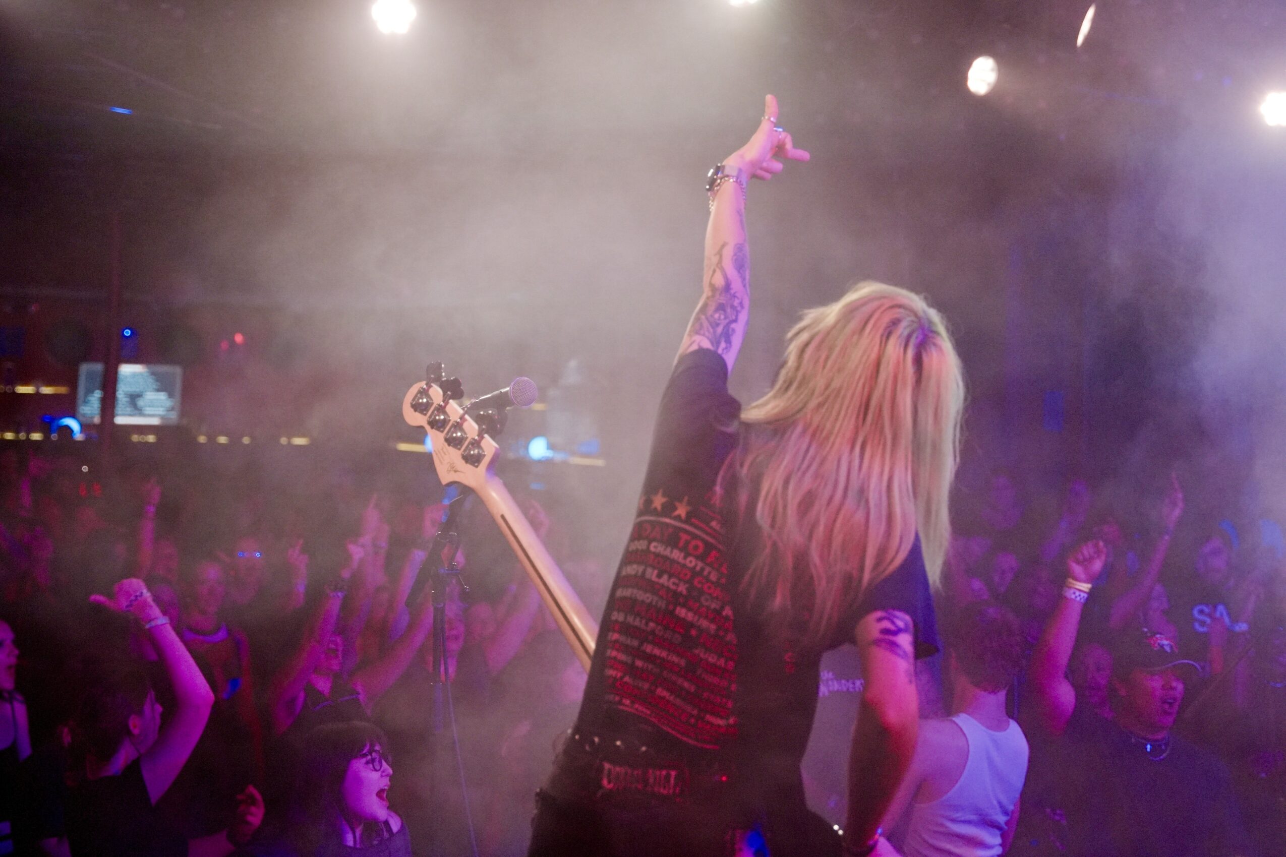 Person with long hair on stage raising one arm, holding a guitar, with a crowd in the background, stage lights, and fog.