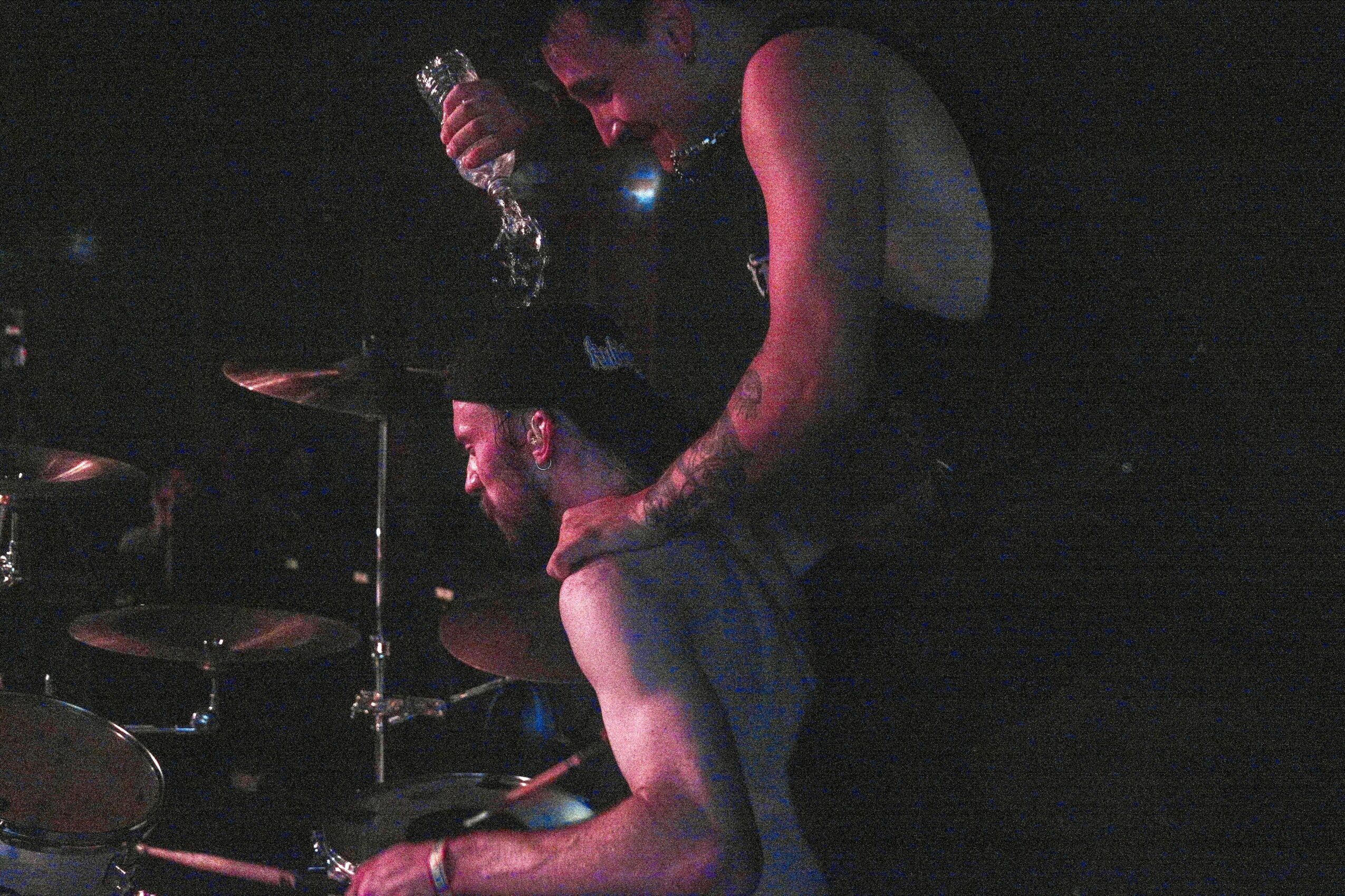 Two people, one standing and one sitting, in a dark setting with musical instruments visible, engaging in an activity.