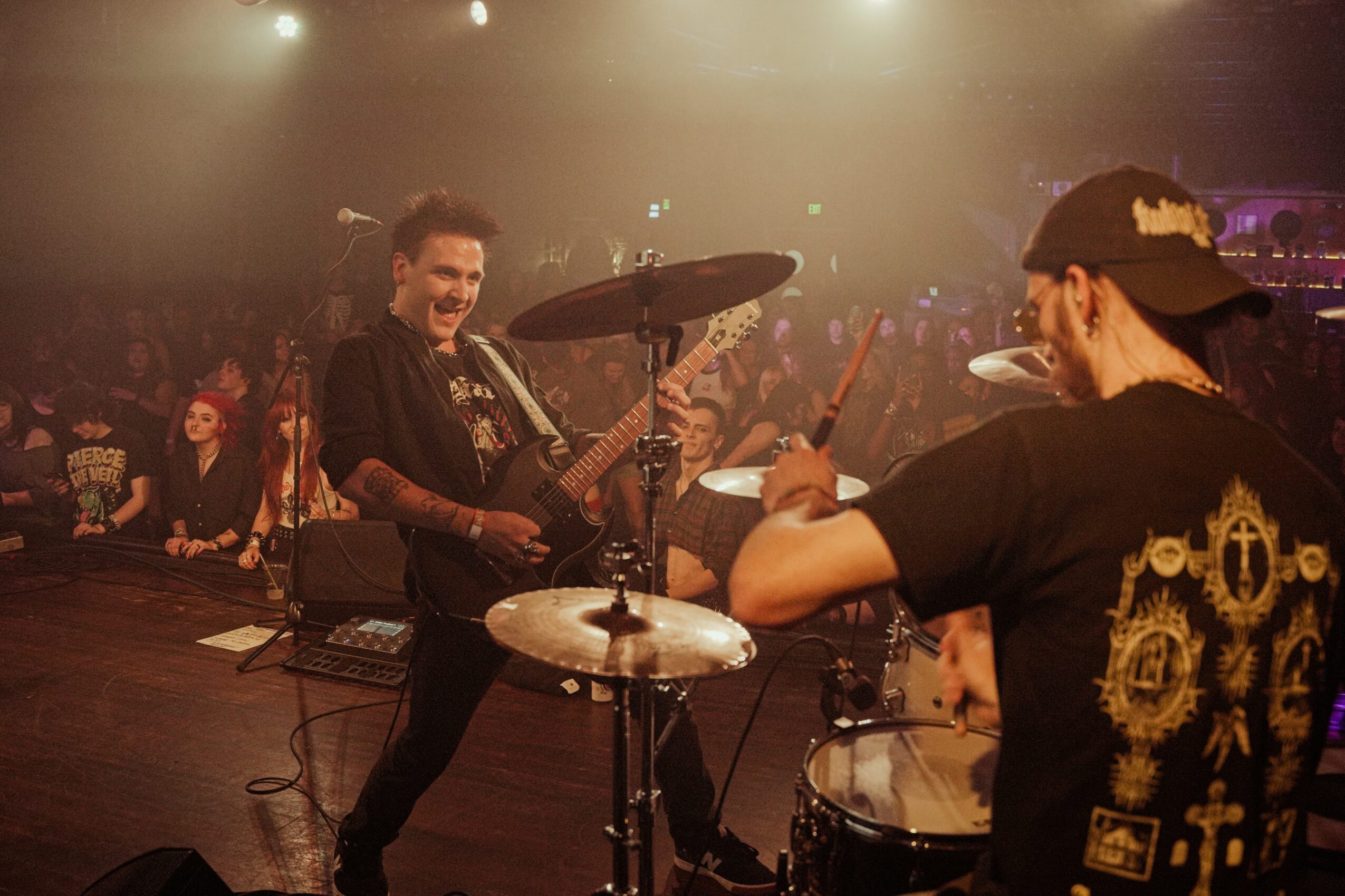 Two musicians perform on stage with audience in background, one playing guitar and the other on drums, under stage lights.