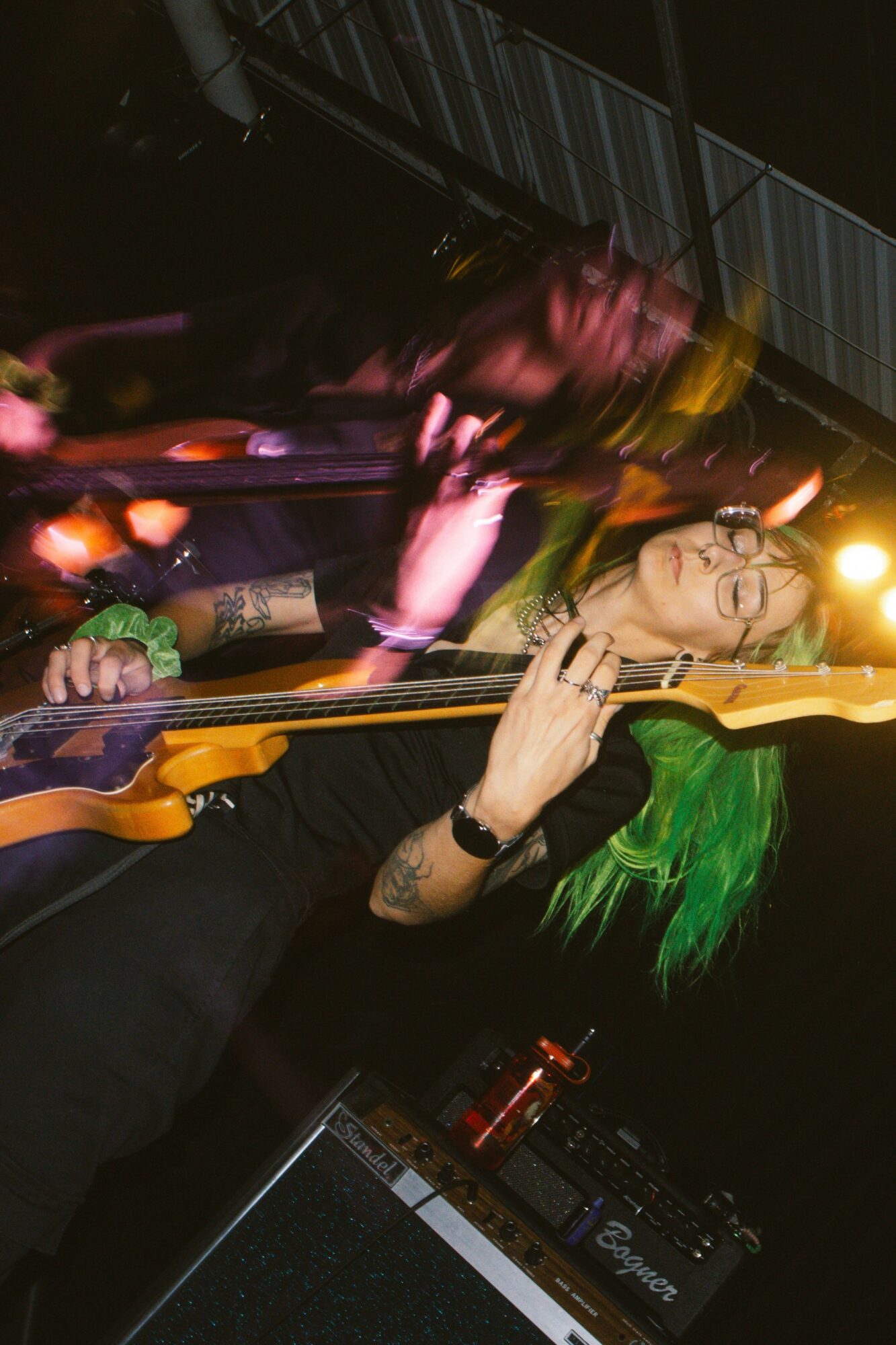 Person with green hair and glasses playing guitar on stage, with colorful lighting and equipment in background.