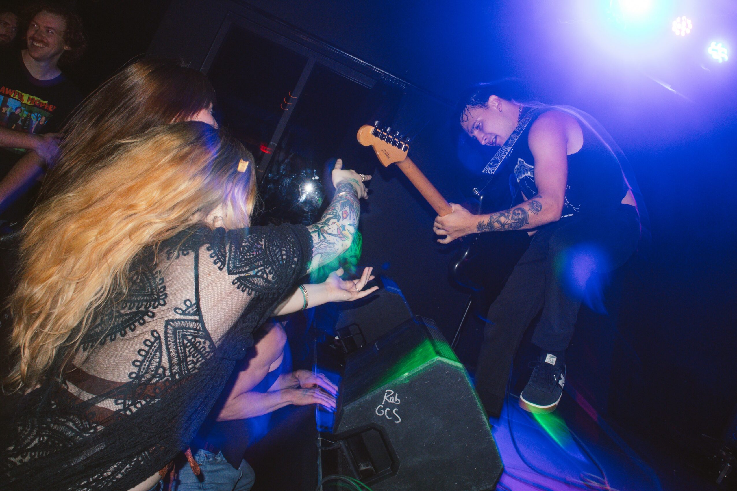 Musicians perform on stage with colorful lighting, one playing guitar and others reaching out to the audience.