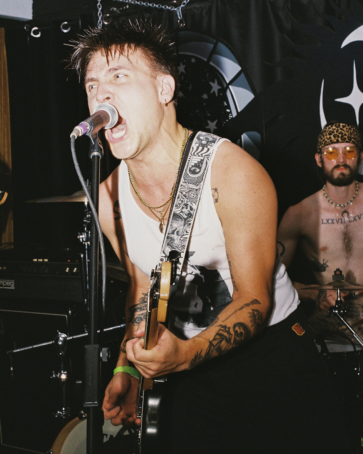 Male musician singing into microphone, playing guitar, with tattooed arm, in sleeveless shirt, and another person in background wearing sunglasses