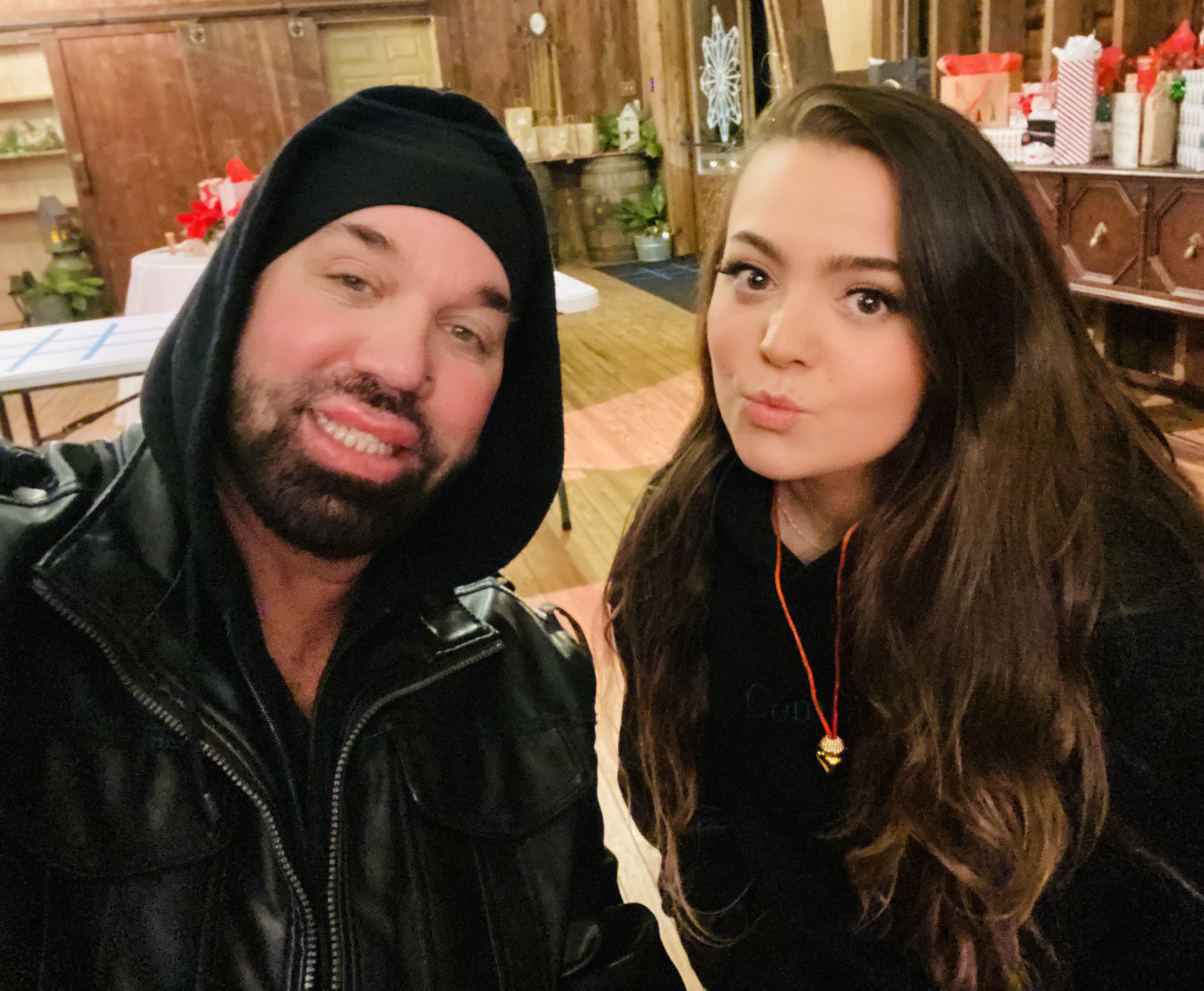 Two people sitting indoors, one wearing a black hoodie and leather jacket, the other with long brown hair and a black top.
