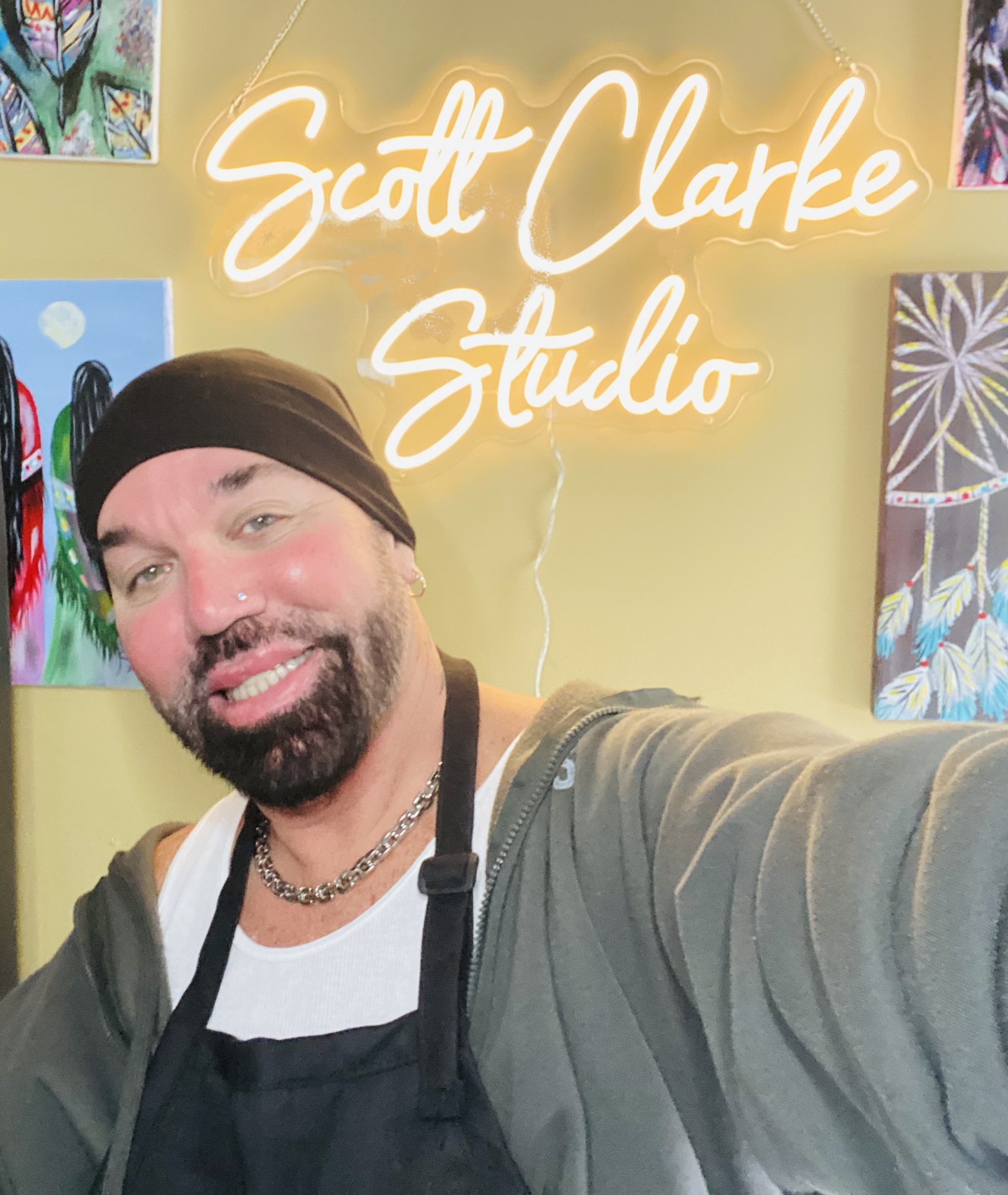 Man with a beard and black beanie taking a selfie in front of a neon sign that says 'Scott Clarke Studio' with colorful artwork on the wall.