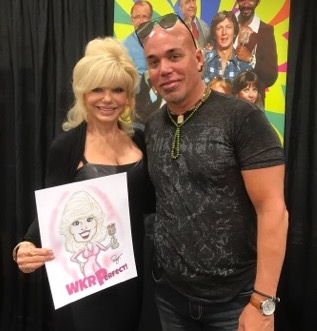 Two people smiling, woman holding a drawing of a girl with pink hair, background with colorful characters, black curtain backdrop.