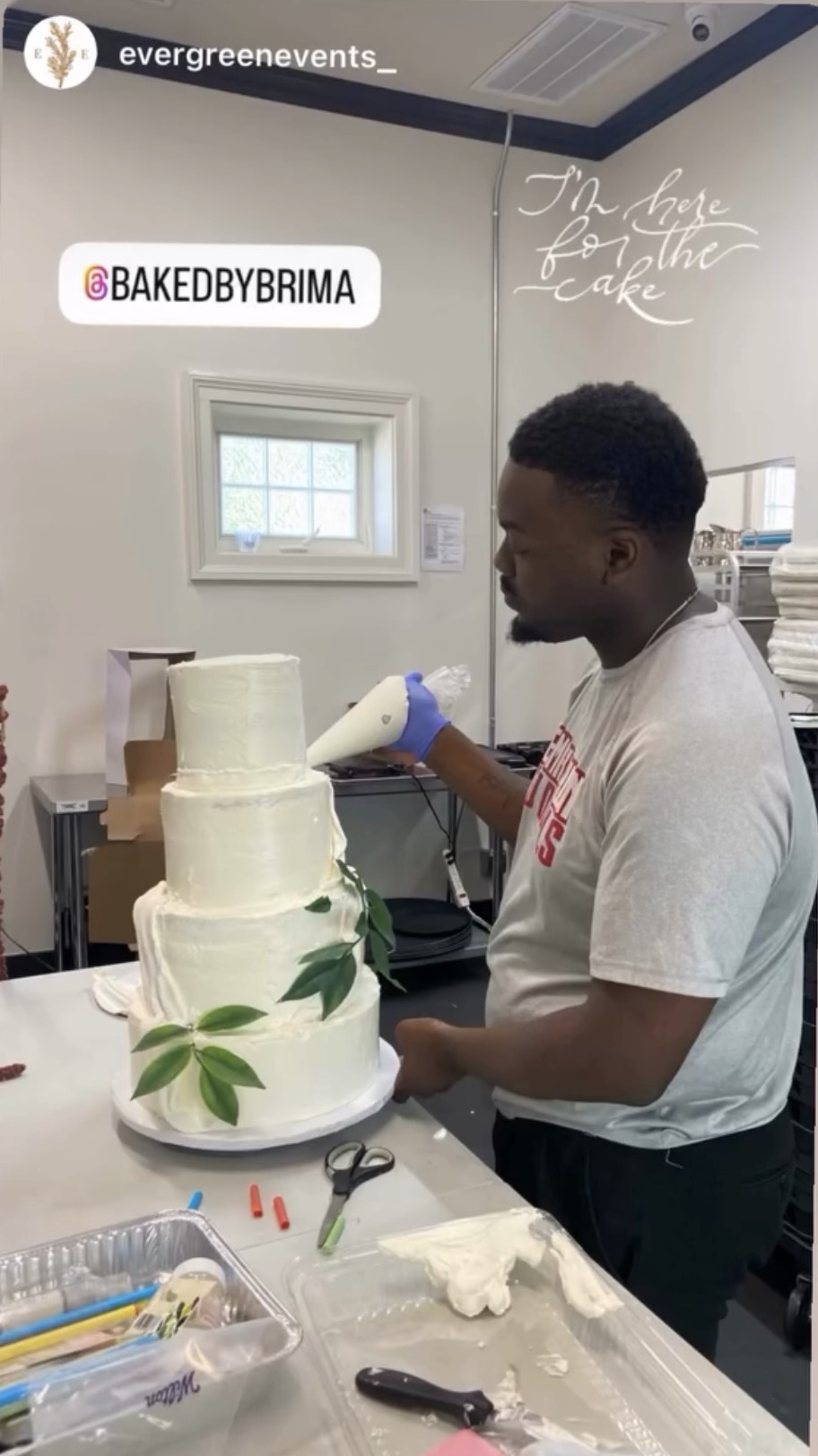 Man decorating a four-tier cake with green leaves in a kitchen, with signs reading 'Baked by Brima' and 'The cake for the love of cake'.