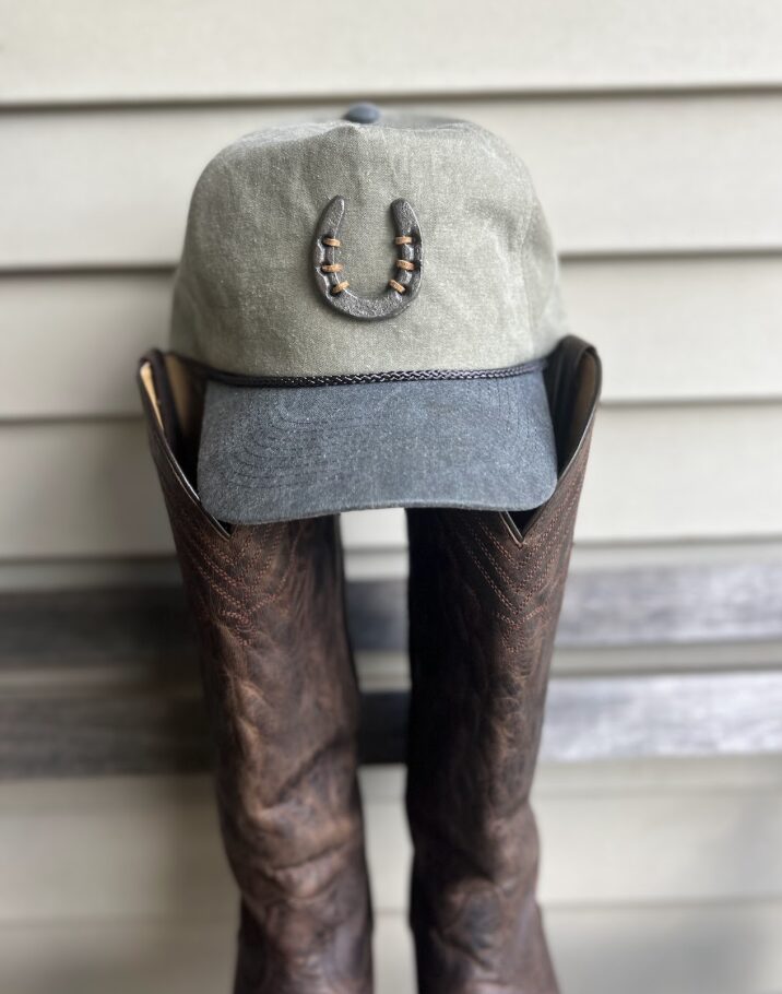 Cap with horseshoe decoration on a stand, with brown leather boots underneath, against a wooden background.