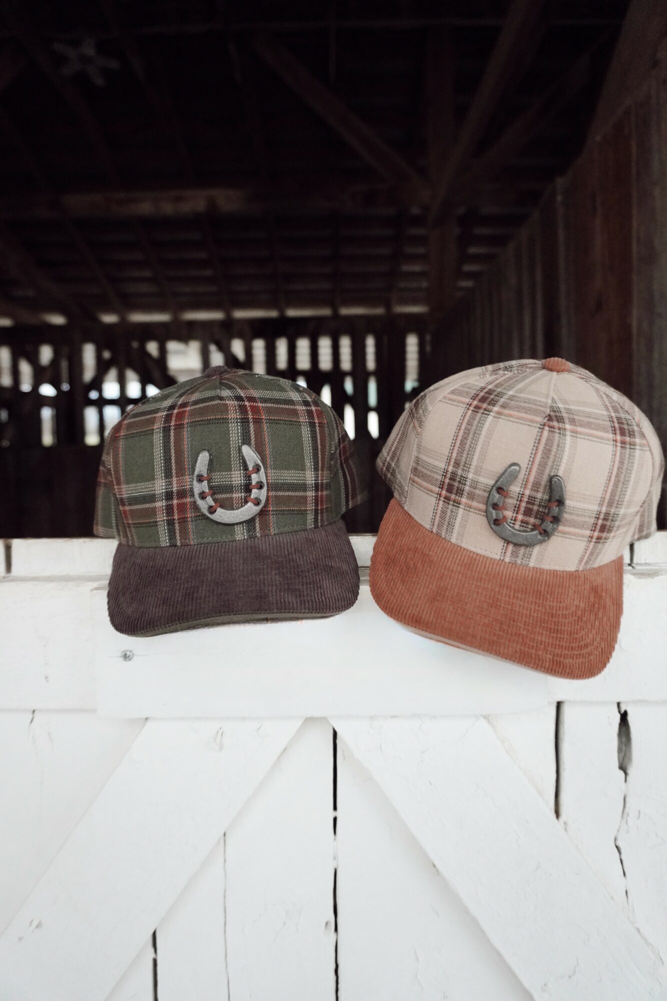 Two plaid caps with horseshoe decorations sit on a white wooden surface inside a barn.