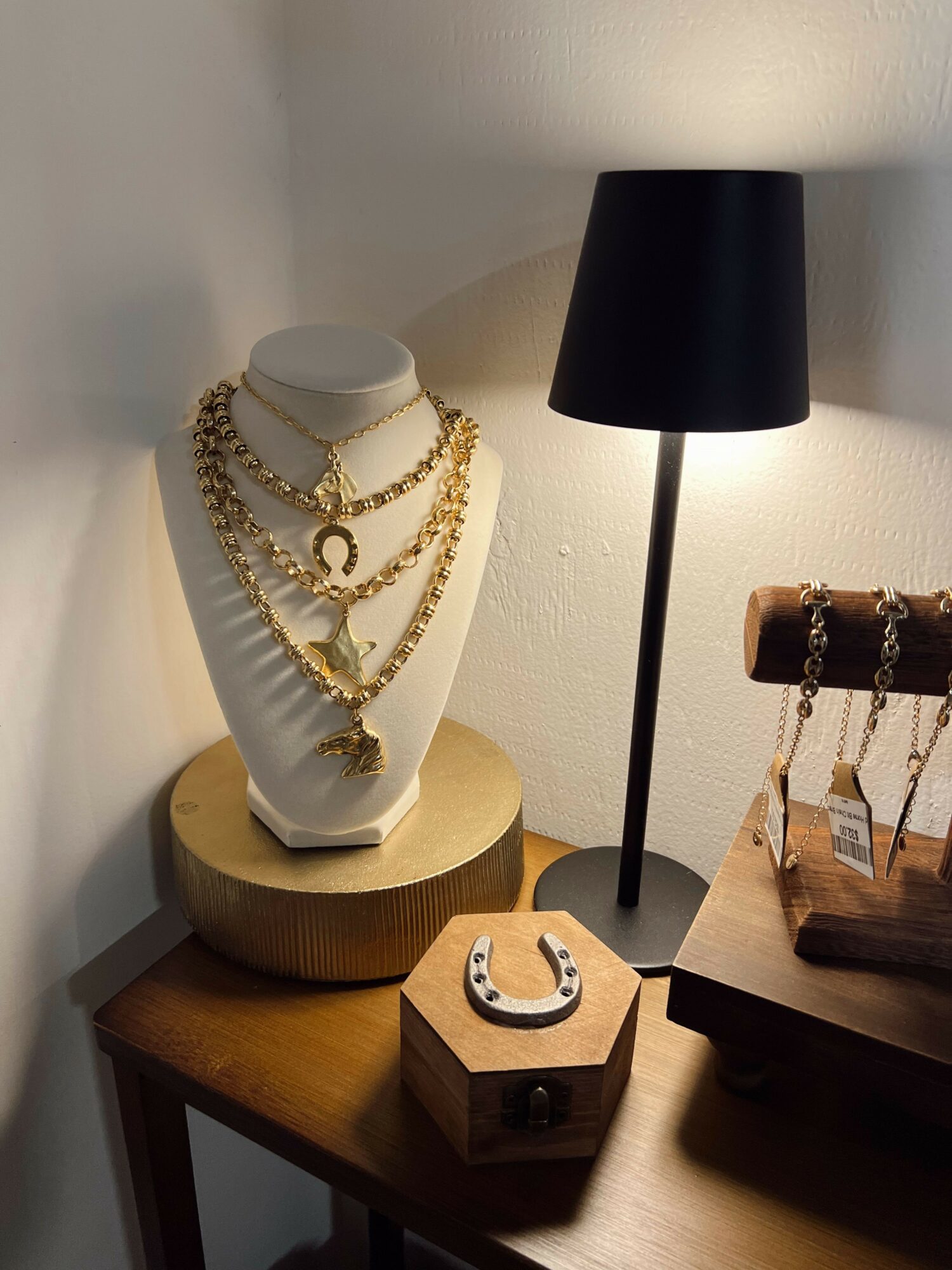 Necklace display on a white bust with a gold chain, on a wooden table with a black lamp and jewelry stand