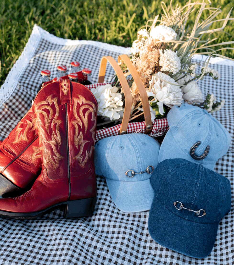 Red cowboy boot, two light blue caps, and a dark blue cap with metal embellishments on a checkered blanket with flowers and grass.