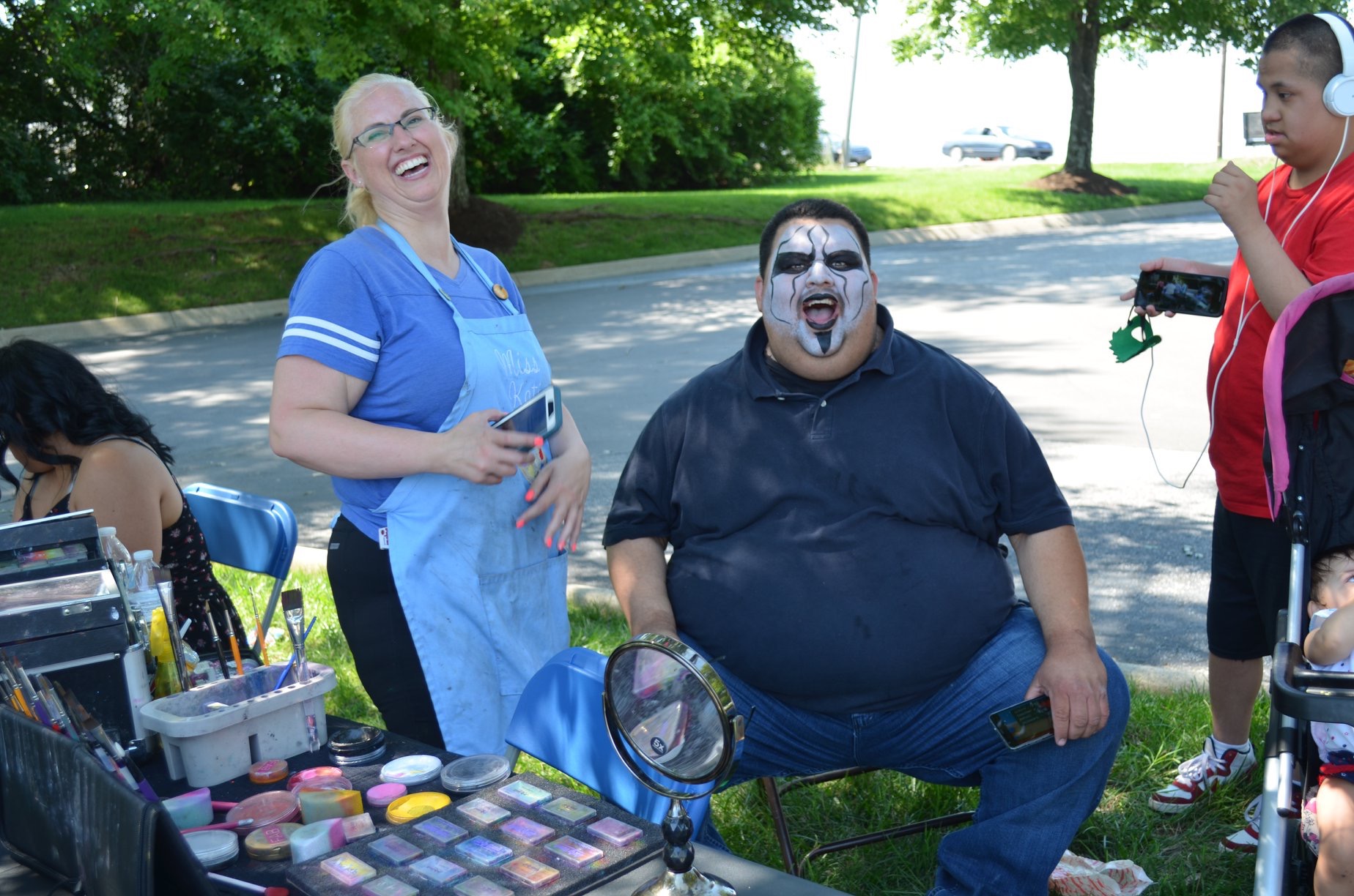 Two people smiling, one with face paint, standing outdoors near makeup and accessories, with children and trees in background.