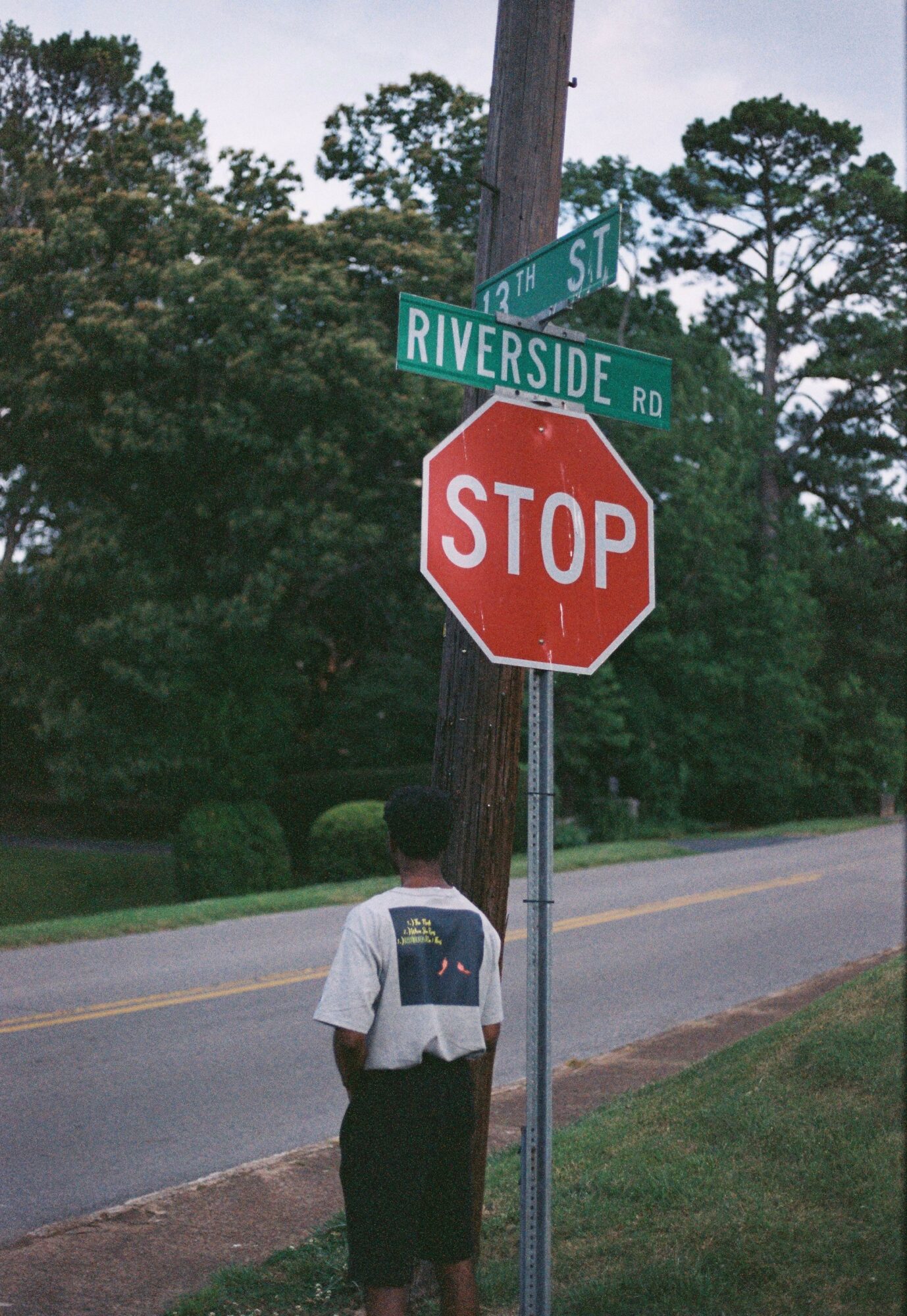 Person standing at corner with stop sign and street signs for Riverside Road and 7th Street, trees in background.