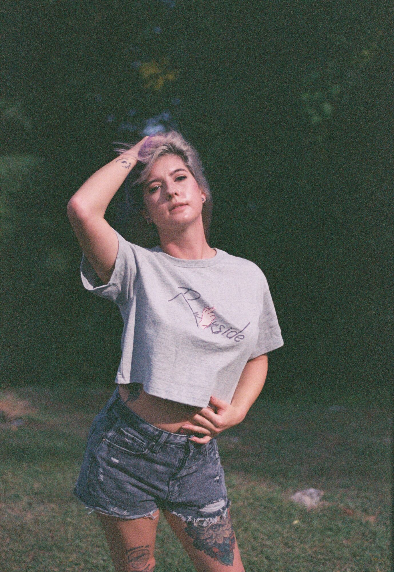 Young woman with short hair wearing a cropped t-shirt and denim shorts, standing outdoors with trees in the background.