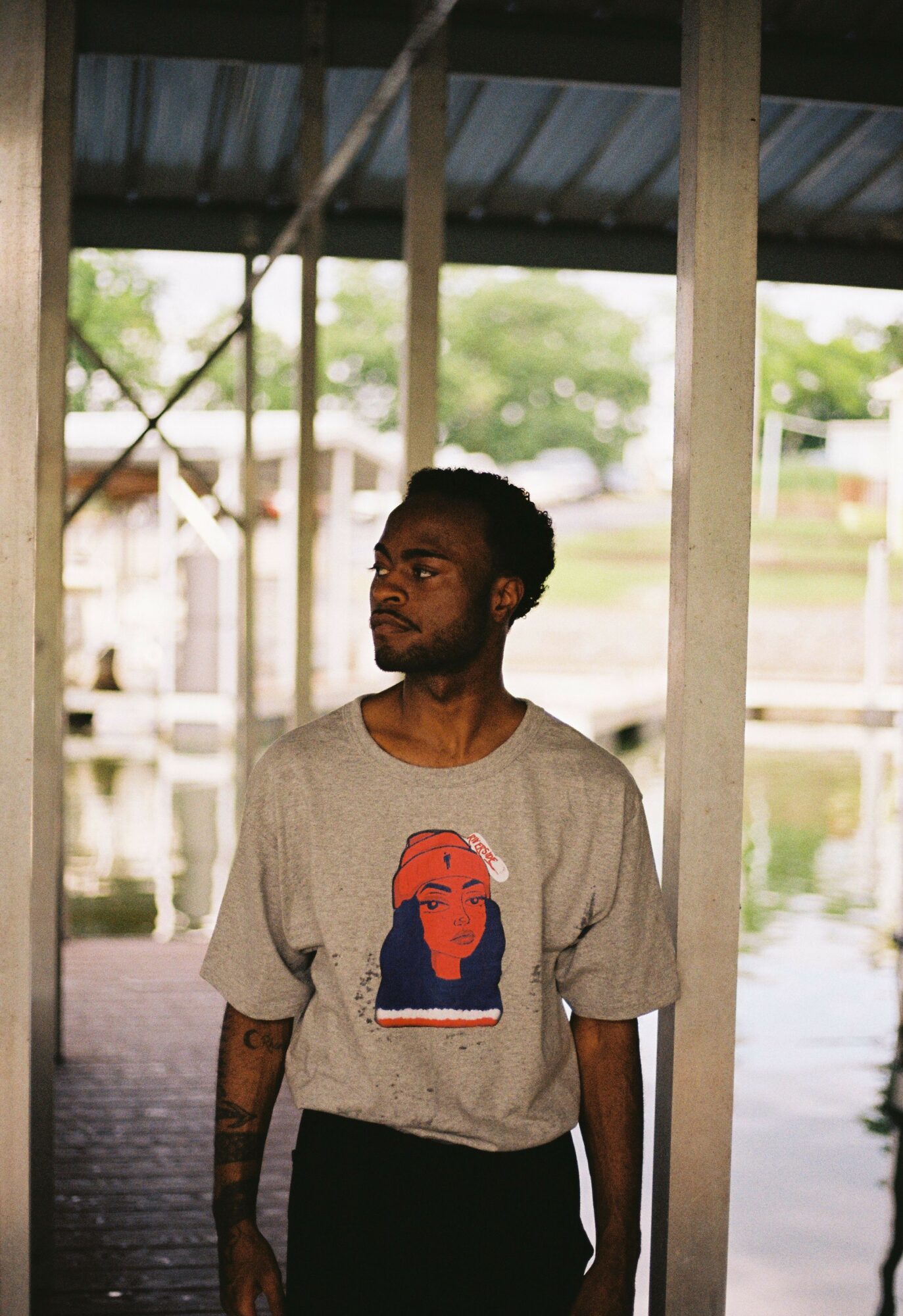 Young man with dark hair and beard standing under a metal roof, looking to the side, near water and trees.
