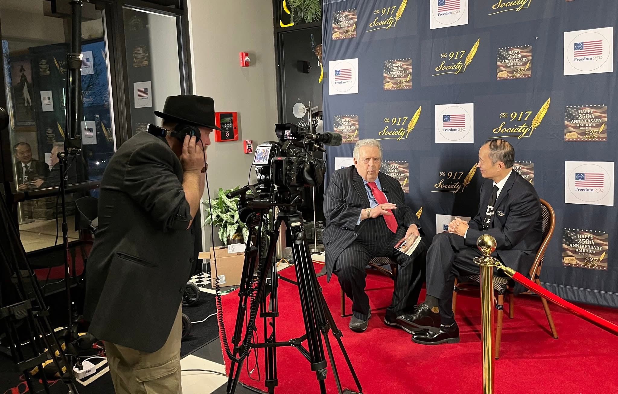 Two men seated on a red carpet, engaged in conversation, with a backdrop and a camera operator recording them.