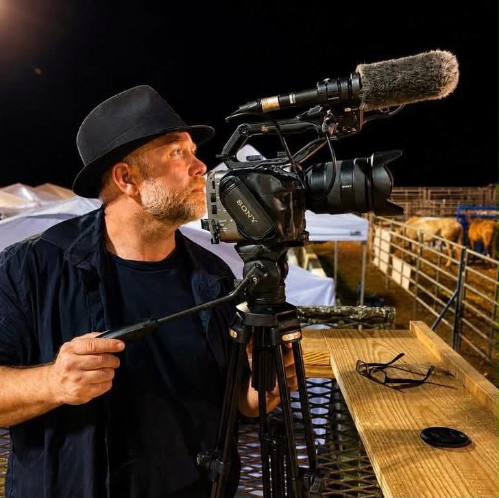 Man operating a professional video camera outdoors at night, with a microphone attached, on a wooden platform.