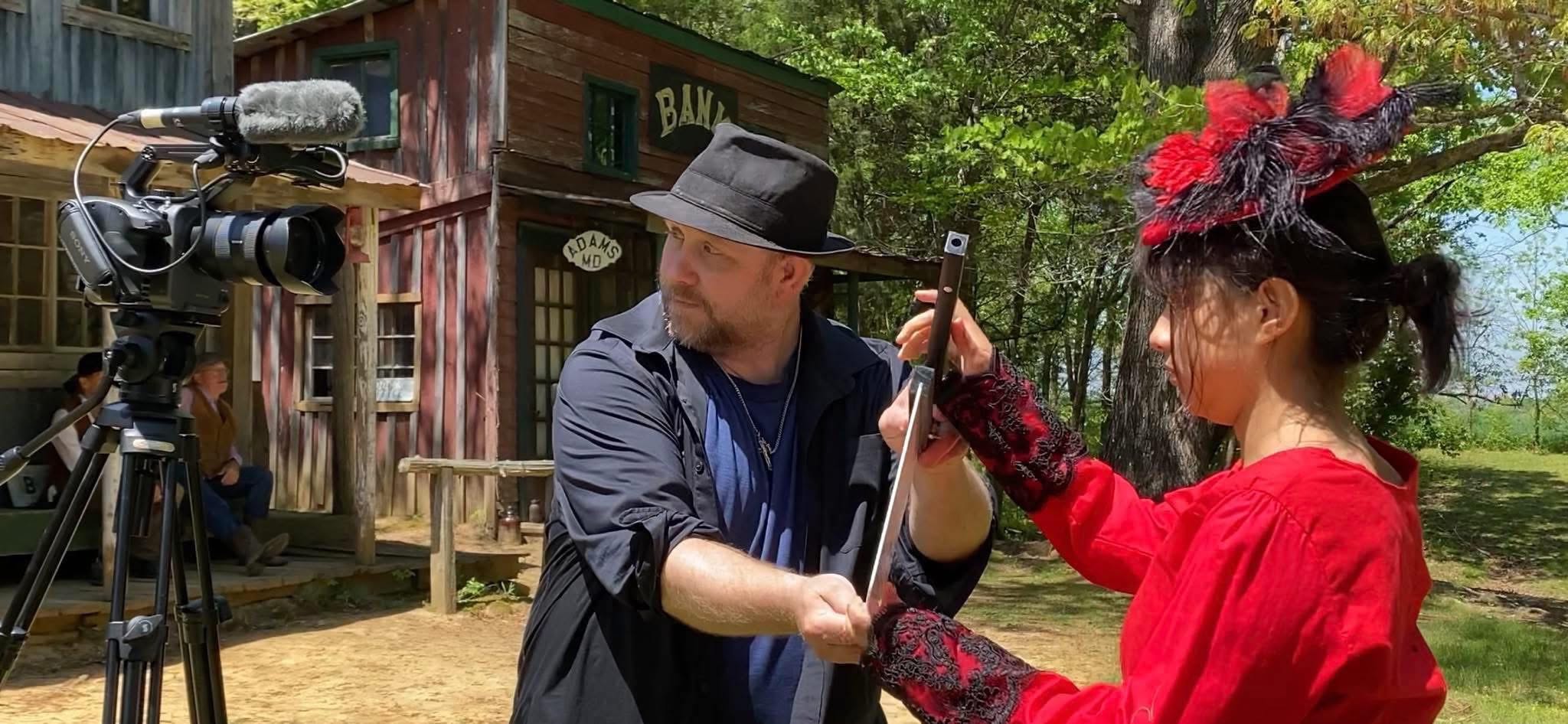 Two people, one in a black hat and the other in a red dress with a hat, are on a film set outdoors.