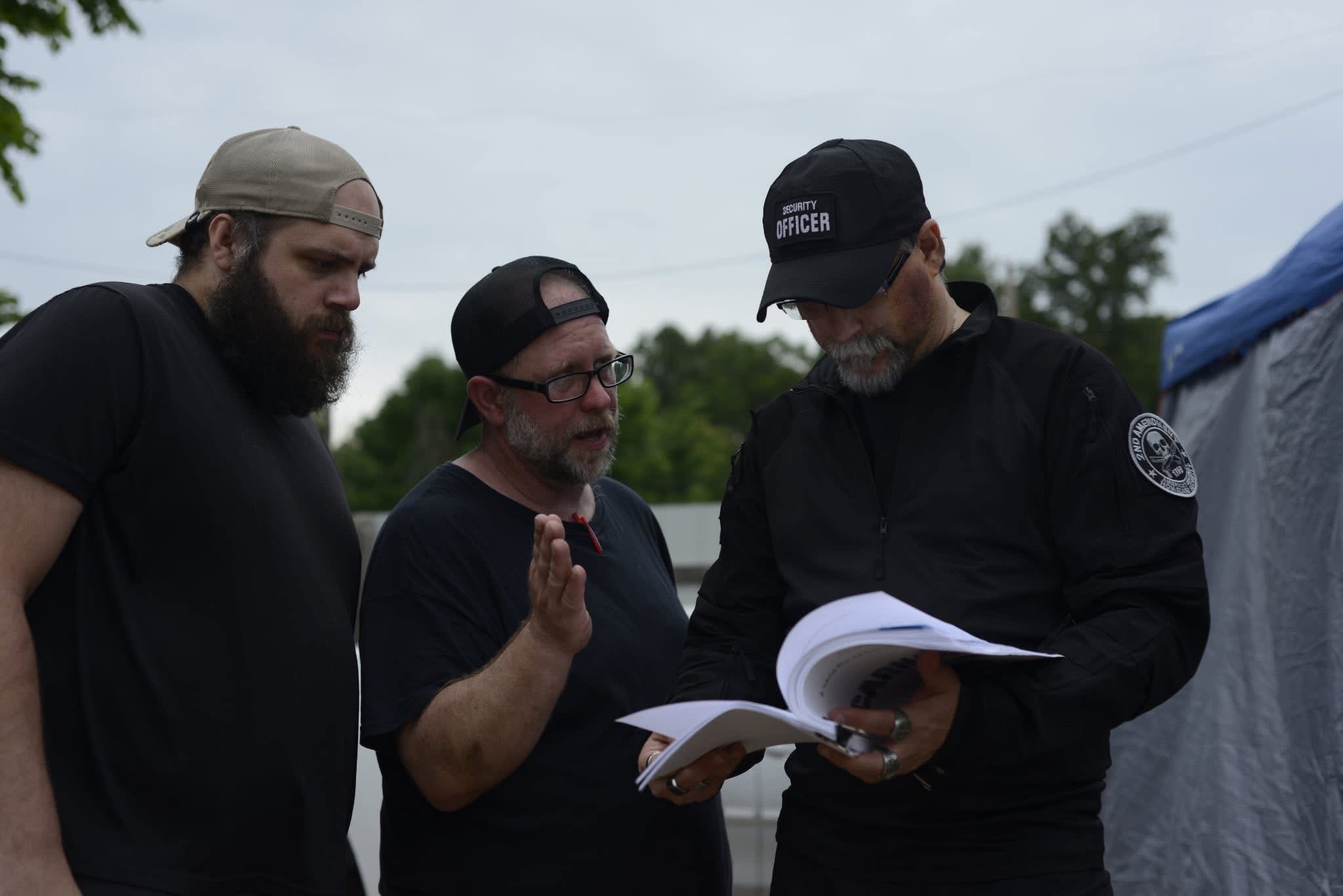 Three men standing outdoors, one holding a book or notebook, engaged in conversation, with trees and a tent in the background.