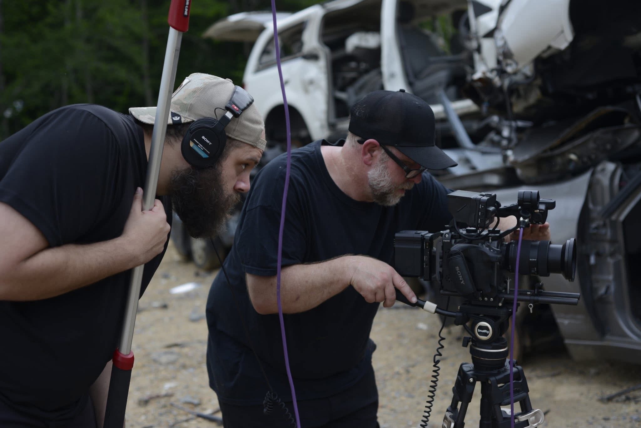 Two men operate a camera on a tripod at a crash site with damaged vehicles in the background.