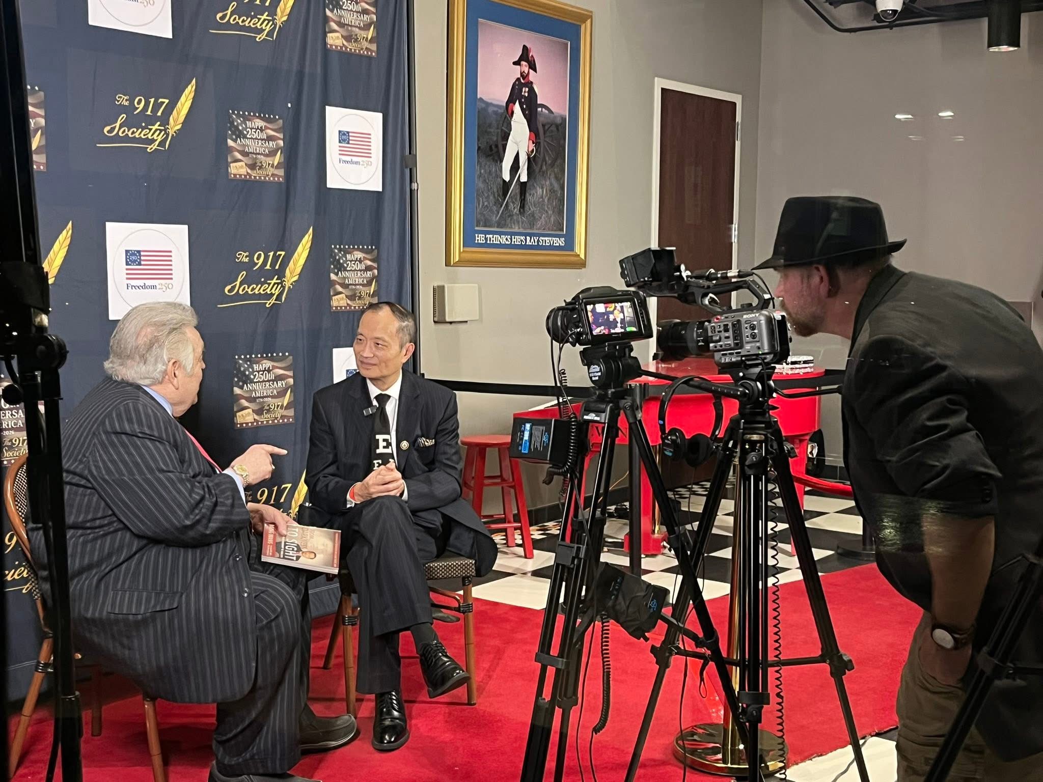 Two men seated and talking in a room with a camera operator filming them, framed pictures on the wall, and red chairs.