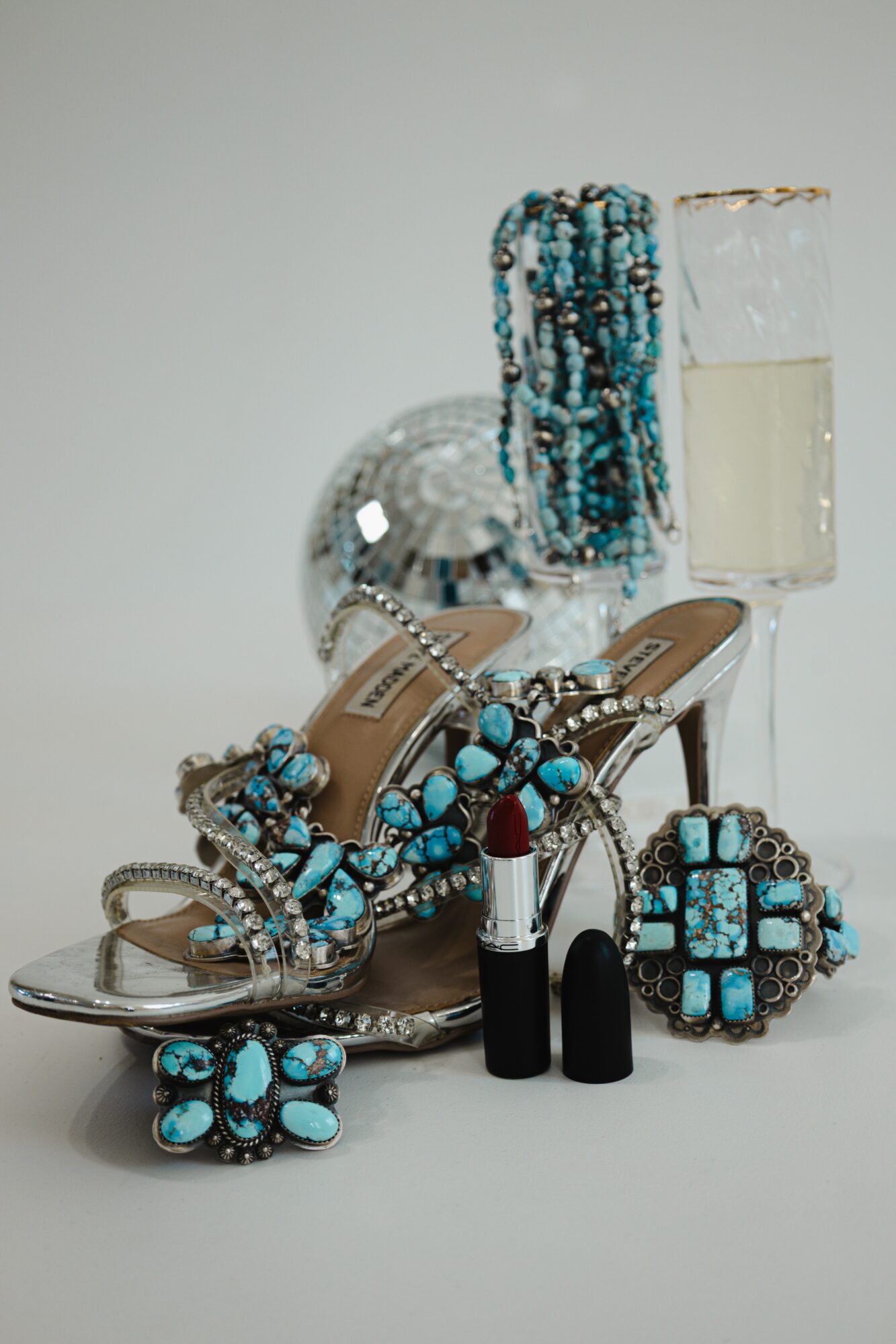 Display of turquoise jewelry, high-heeled shoes, lipstick, and a glass of champagne on a white surface.