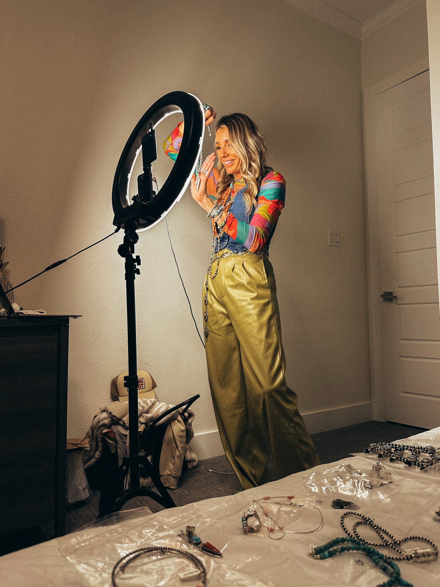 Woman with colorful long-sleeve top and high-waisted pants stands near a ring light in a room, smiling and looking down.