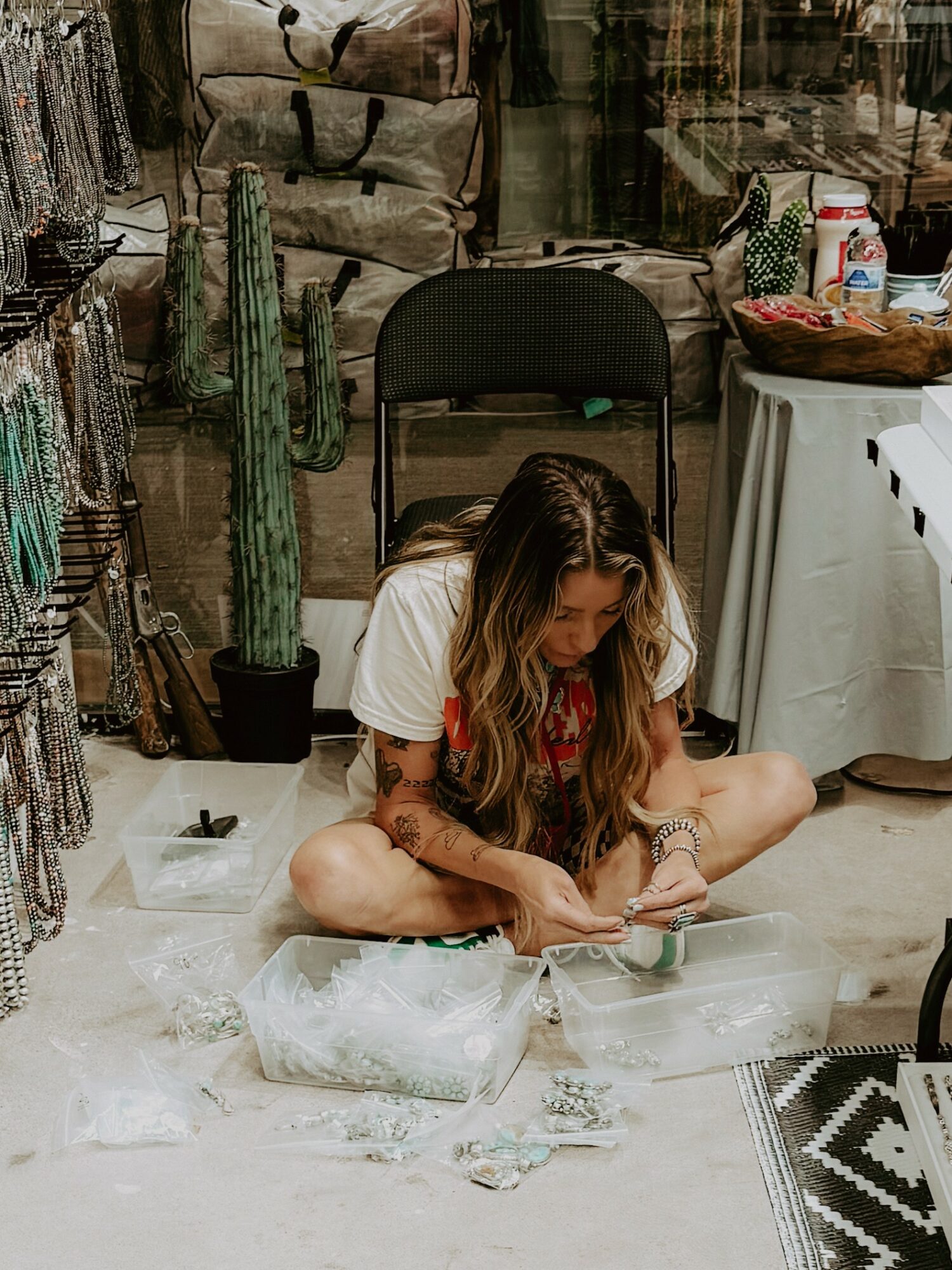 Person sitting on the floor with long hair, surrounded by various objects, in a room with plants and furniture.