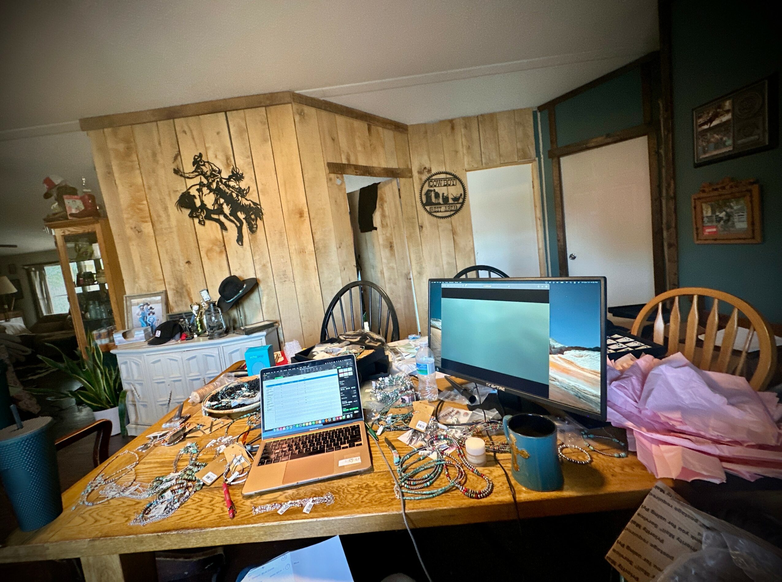 Cluttered dining table with two laptops, a monitor, and various items in a room with wooden walls and chairs.