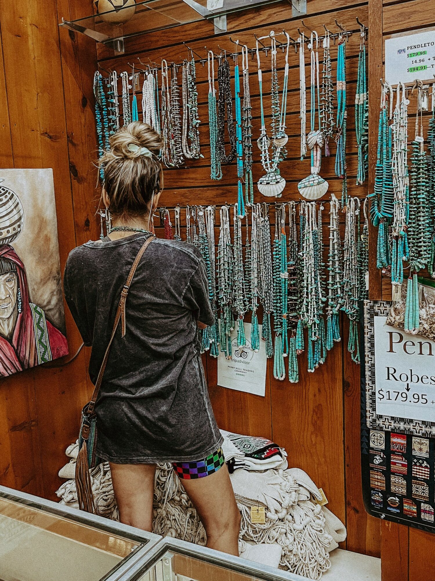 Person with a bun hairstyle wearing a dark jacket shopping for necklaces at a jewelry display, with wooden wall background.