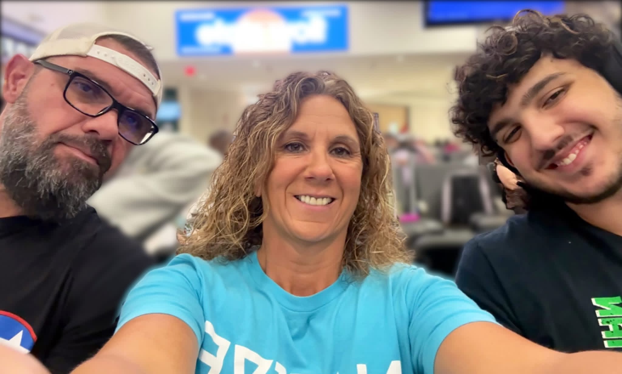Three people smiling in an indoor setting, with a blurred background and a blue sign above them.