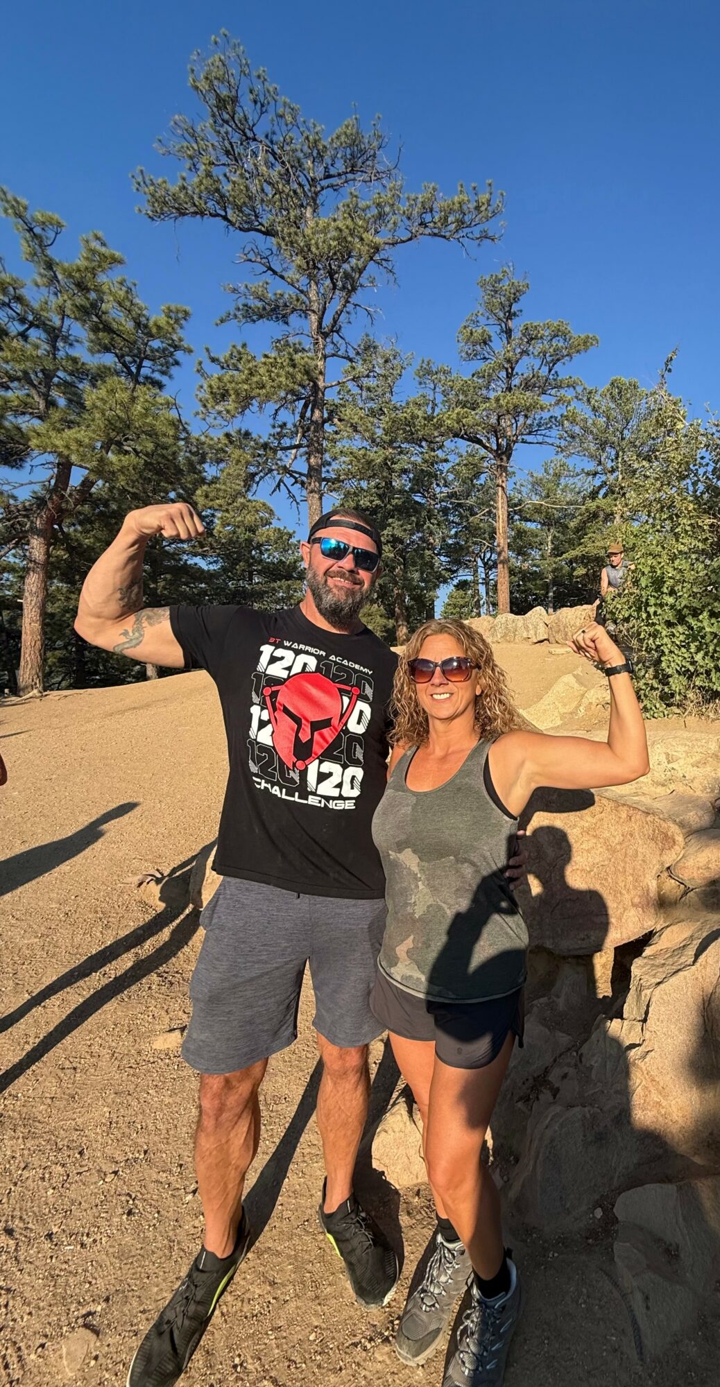 A man and woman flexing their muscles outdoors with trees and blue sky in background.