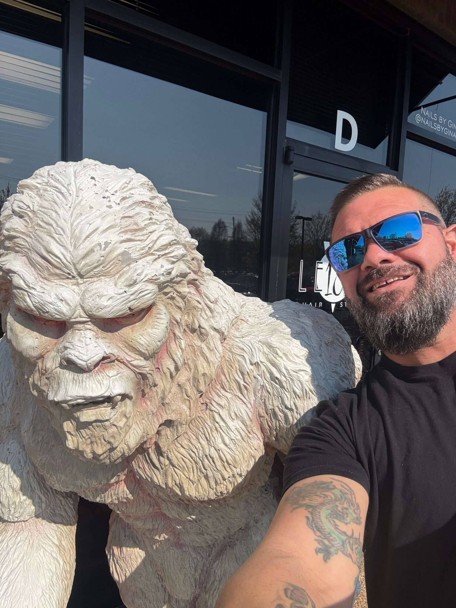 Man with sunglasses and a tattoo takes a selfie with a large, light-colored dog outside a building.