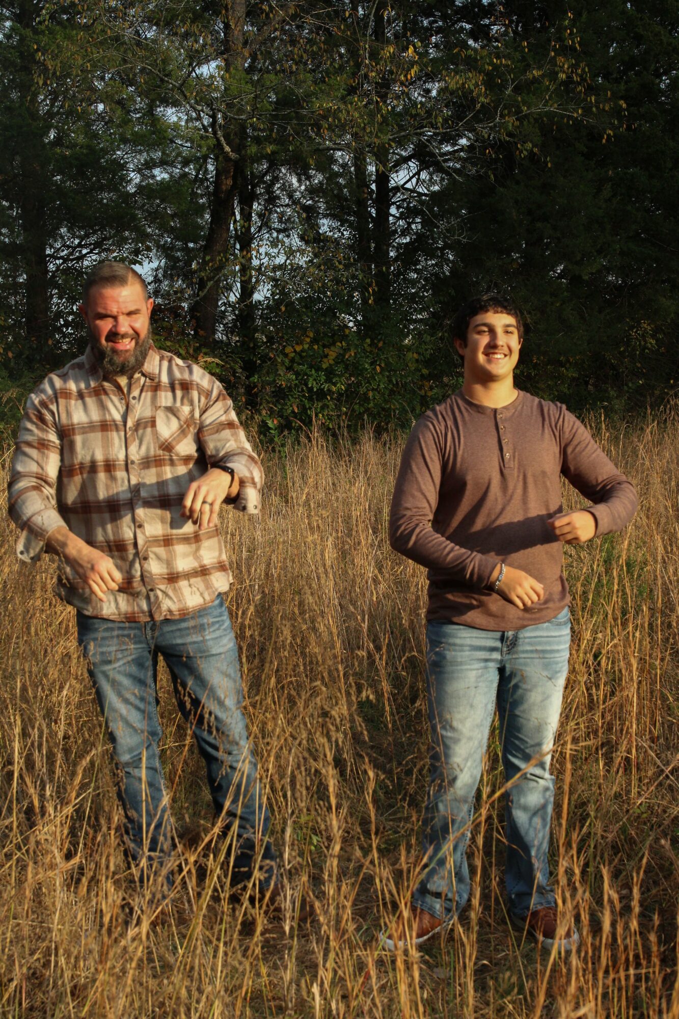 Two men standing in a field with tall grass and trees in the background, smiling and looking at the camera.