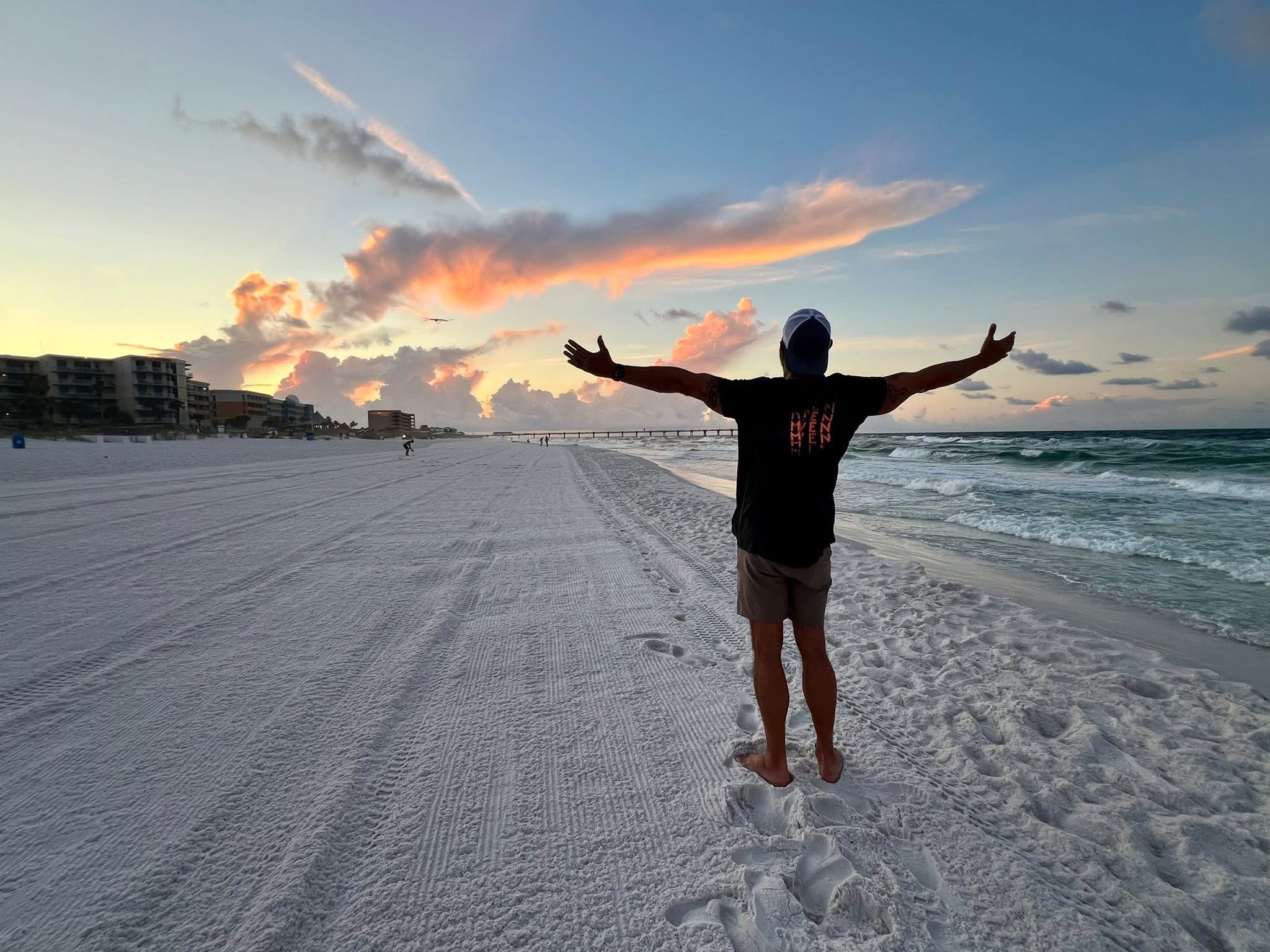 Person standing on beach with arms outstretched, facing the ocean during sunset, clouds in sky, footprints in sand