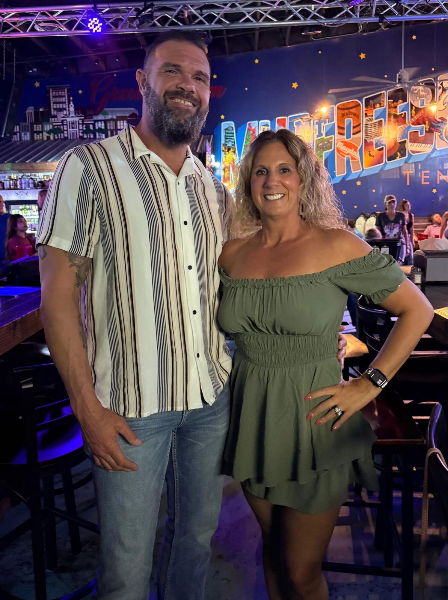 Two people, a man and a woman, stand together smiling in a colorful indoor setting with a stage and lights behind them.