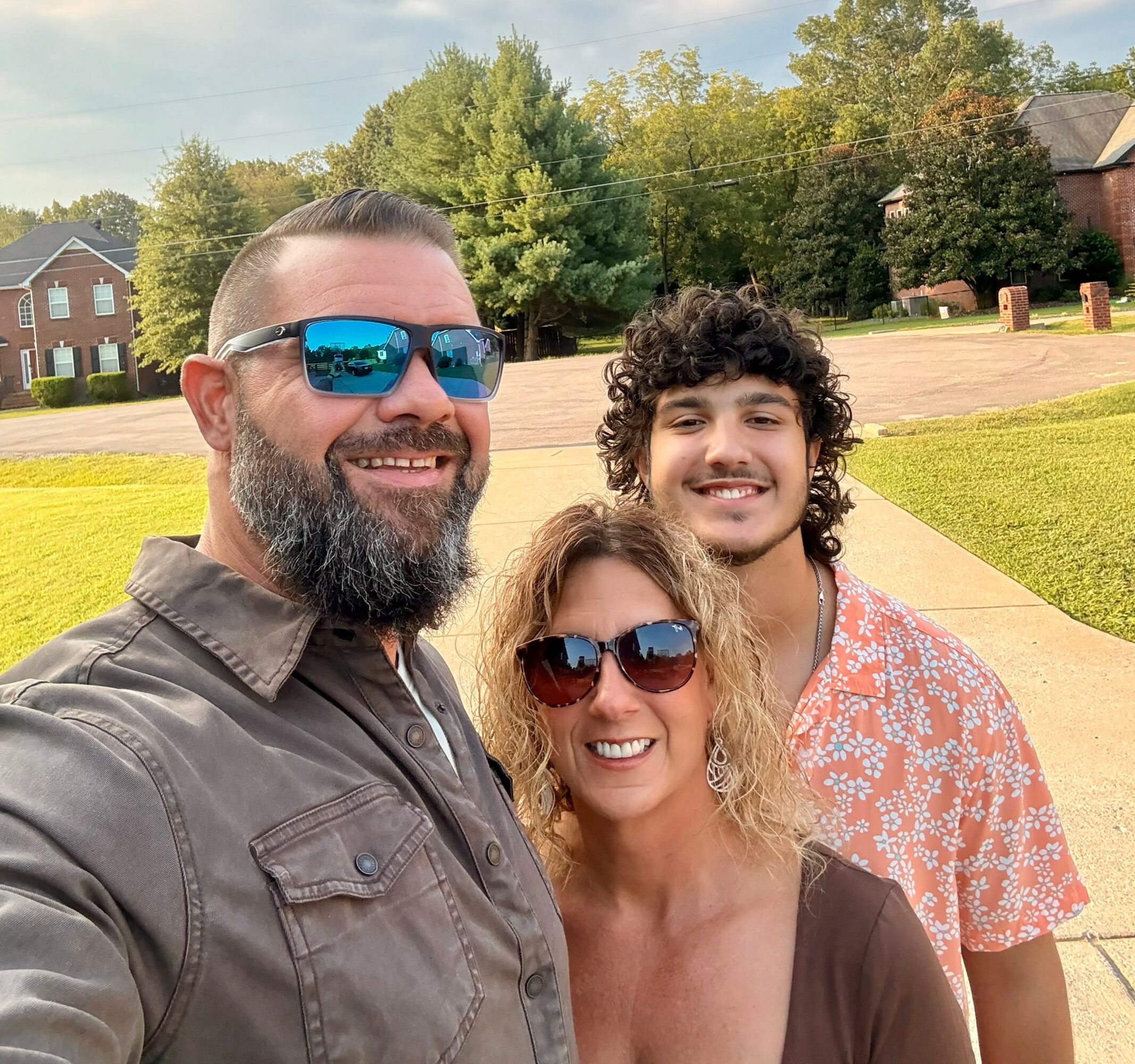 Three people smiling outdoors, two wearing sunglasses, with trees and houses in the background.