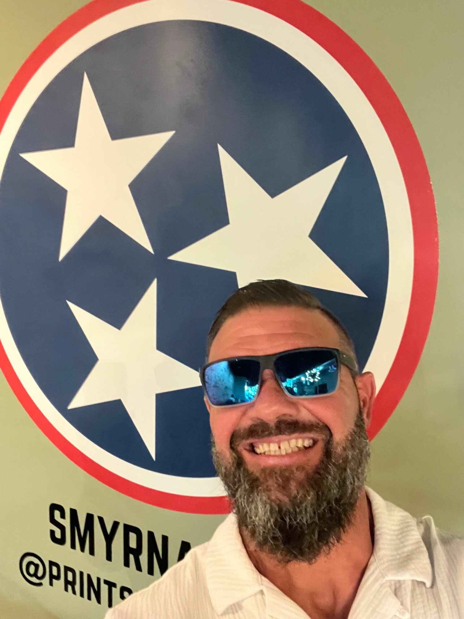 Man with sunglasses smiling, standing next to a Tennessee flag with three stars, partially visible text, and a red, white, and blue background.