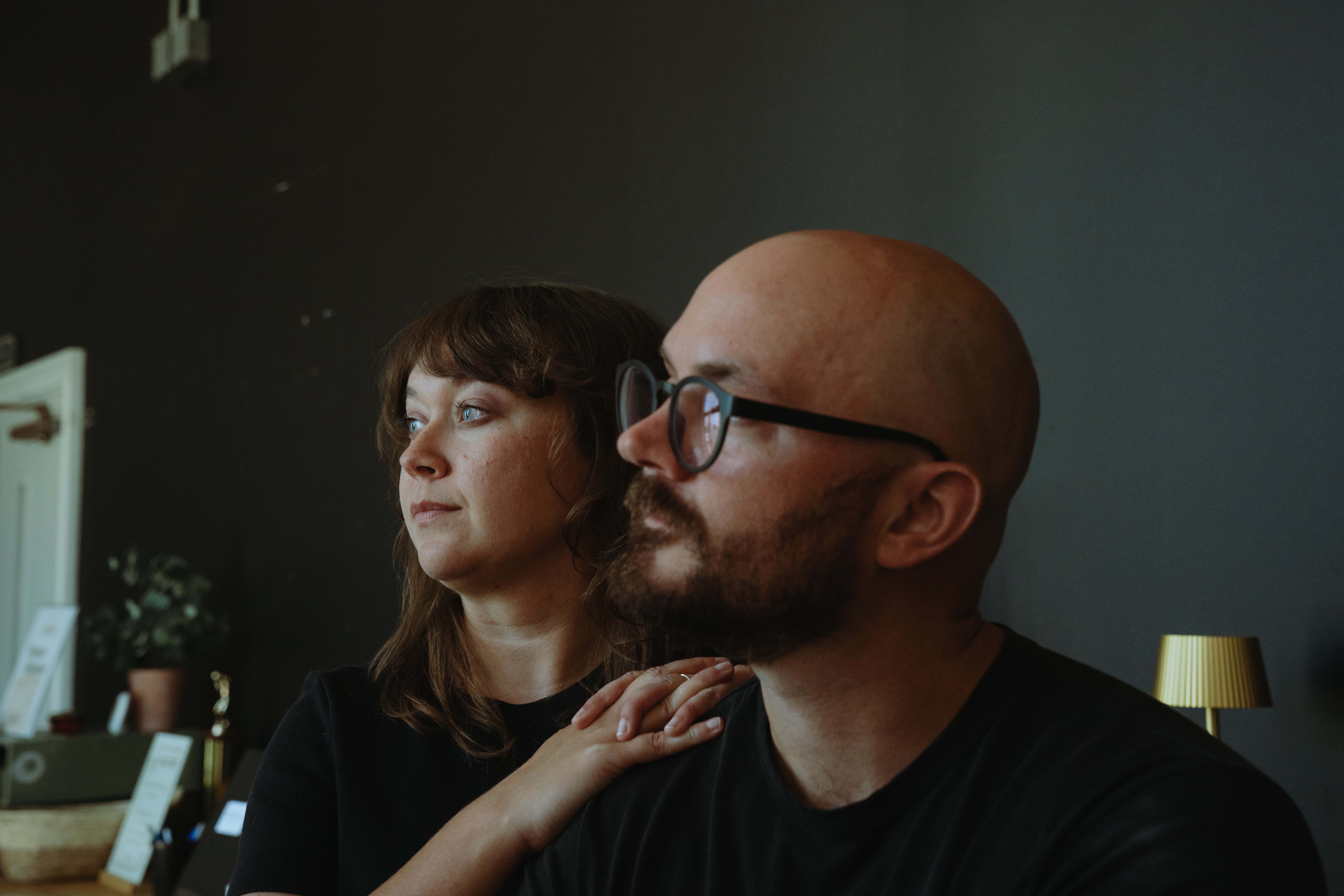 Two people sit close together indoors, one with glasses and a beard, the other with shoulder-length hair, both looking to the side.