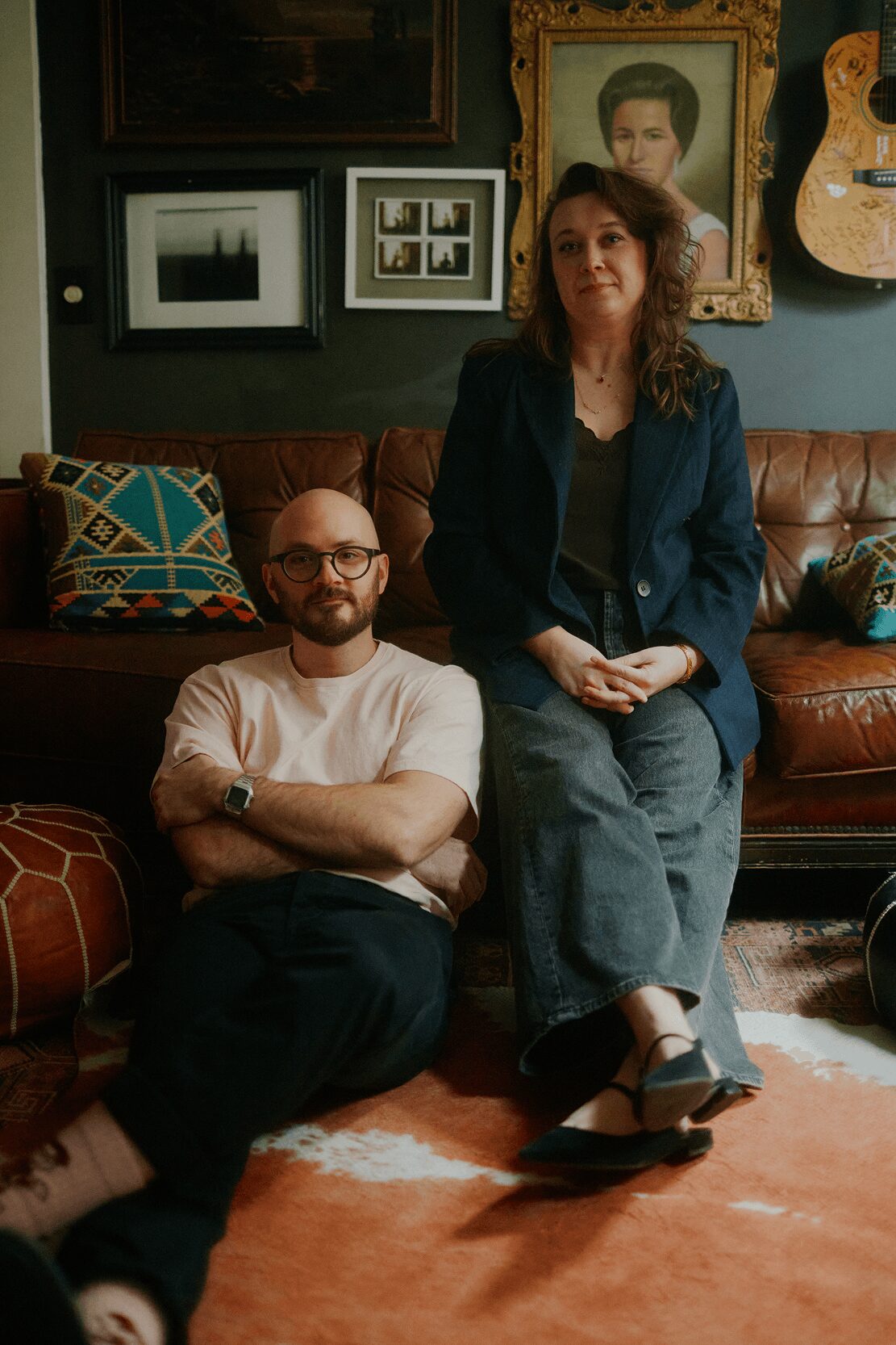 A man with glasses and a woman with curly hair sit and stand in a living room with framed pictures and a guitar on the wall.