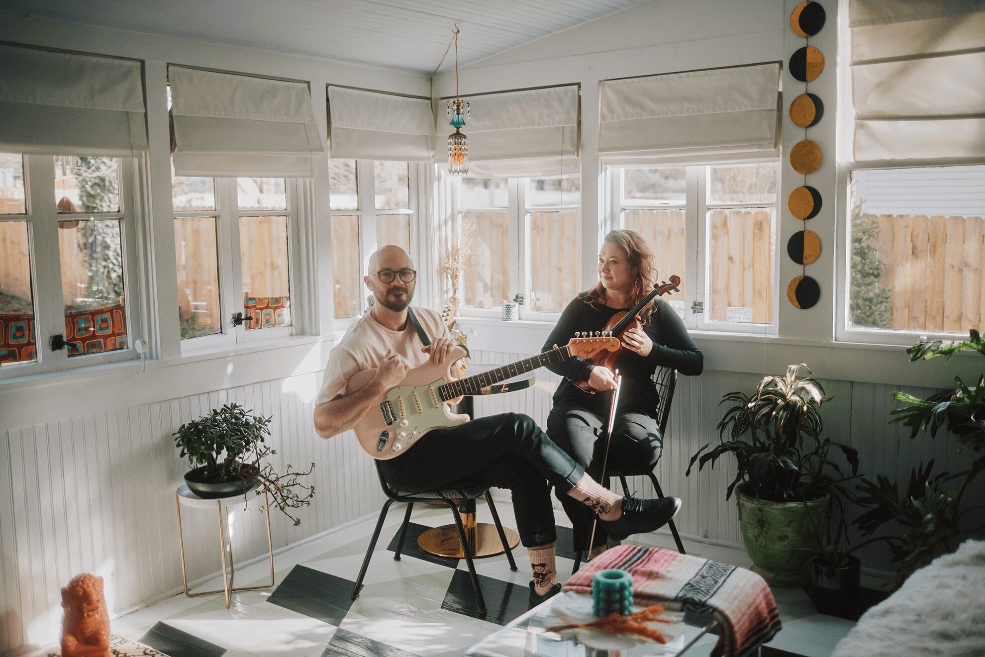 Two people playing guitars in a sunlit room with large windows, plants, and a colorful tablecloth, smiling and enjoying music.