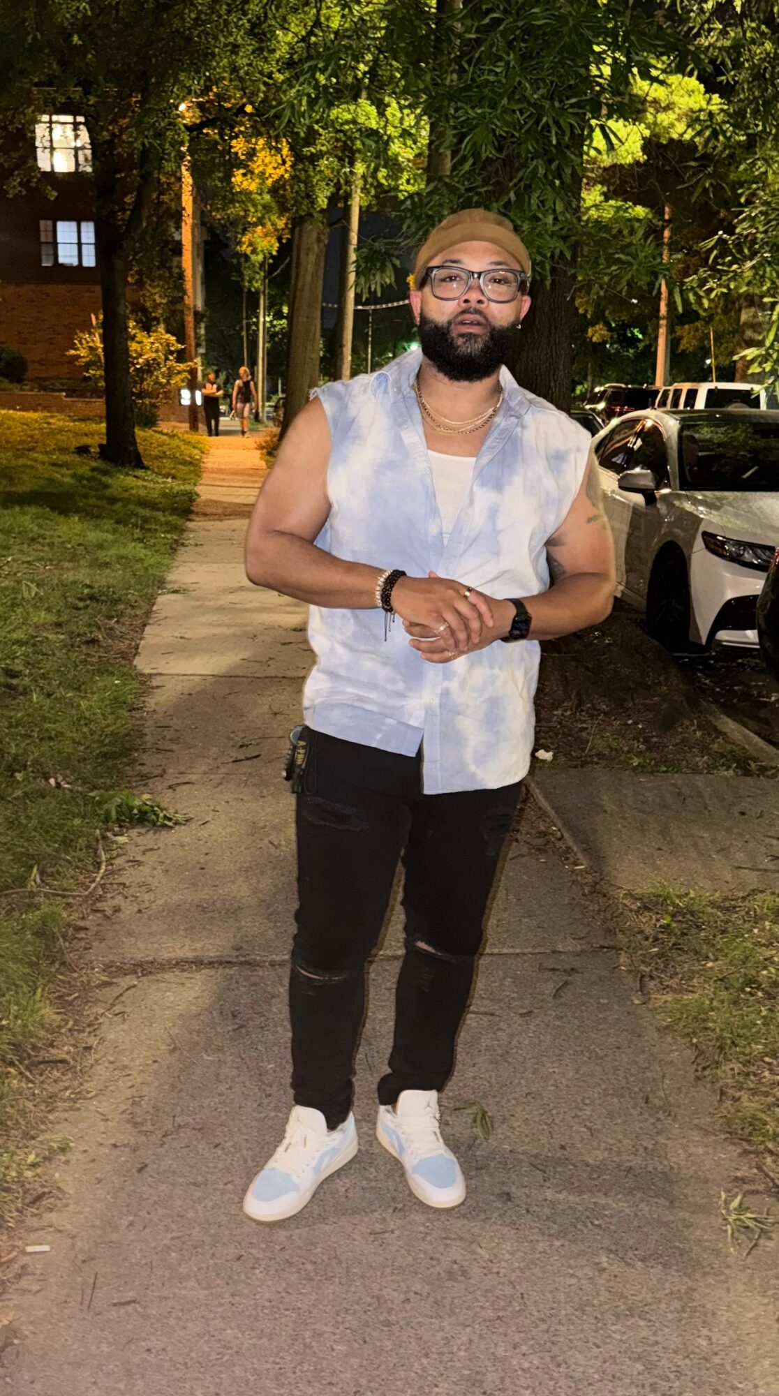 Man with glasses and beard standing on sidewalk at night, smiling, wearing sleeveless denim shirt, black pants, and white sneakers.