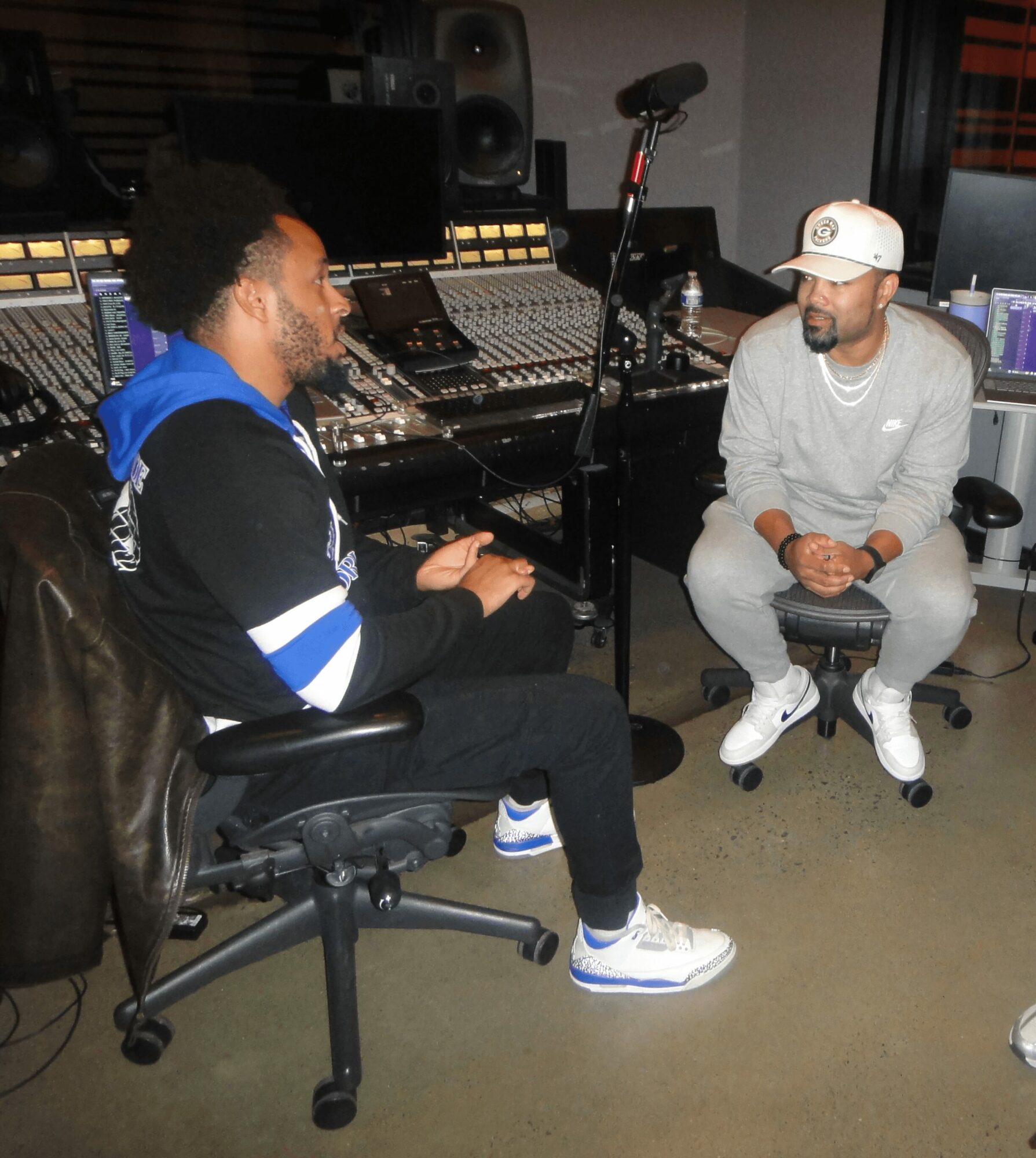 Two men sit in a recording studio with audio equipment and mixing consoles, engaged in conversation. One wears a black hoodie and jeans, the other a gray sweatshirt and cap.
