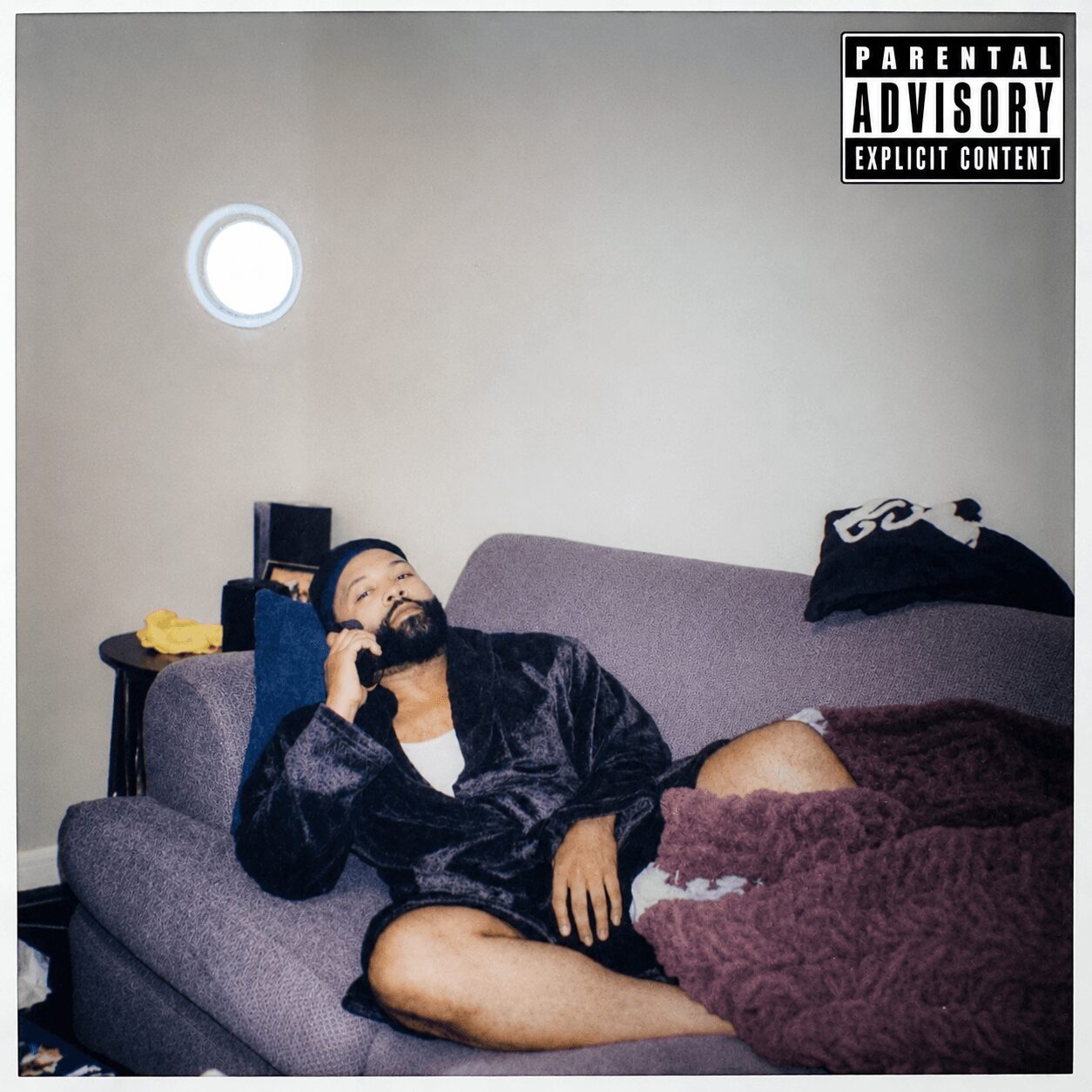 Person lying on a couch, talking on a phone, with a blanket covering their legs, in a room with a plain wall.