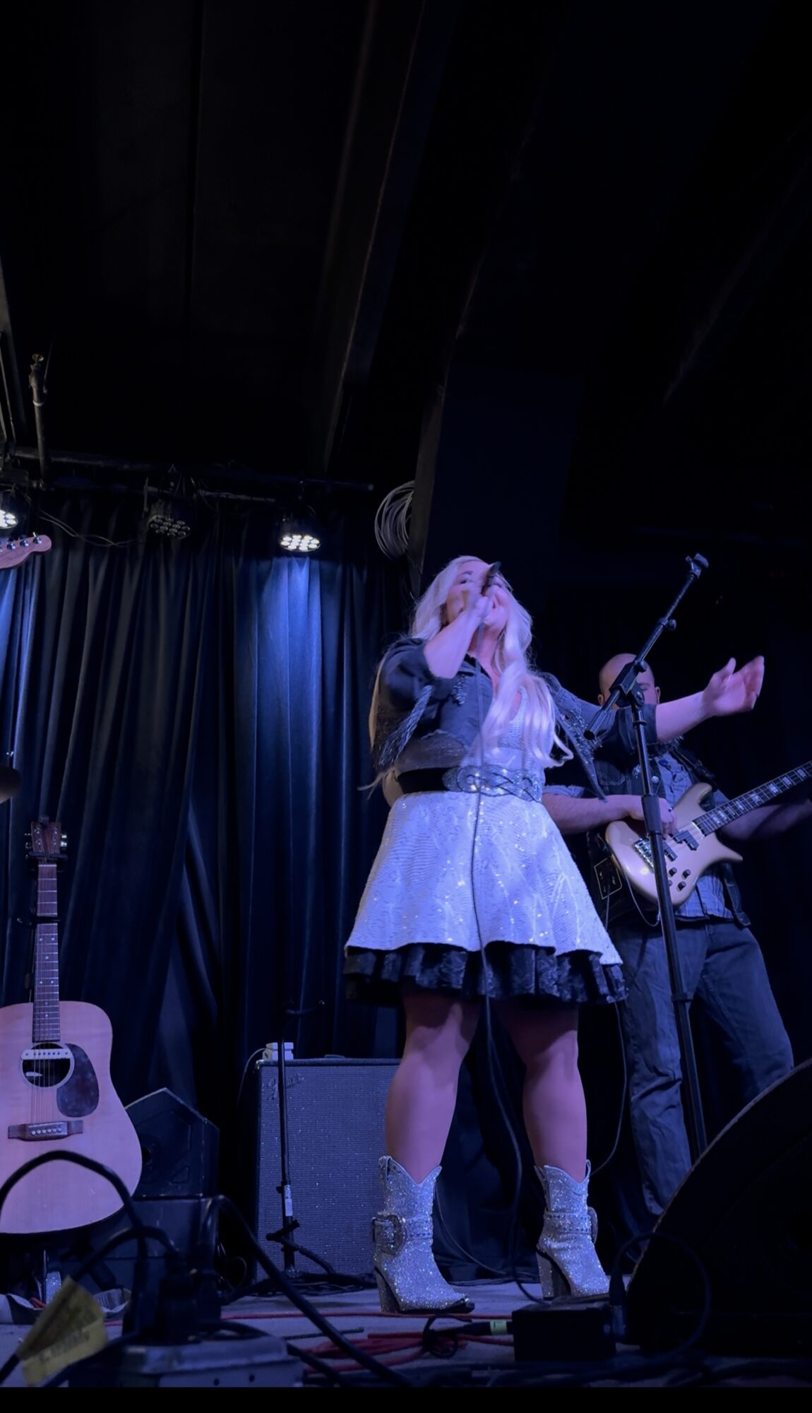 A woman singing on stage with a microphone, accompanied by a guitarist, with guitars and stage equipment visible.
